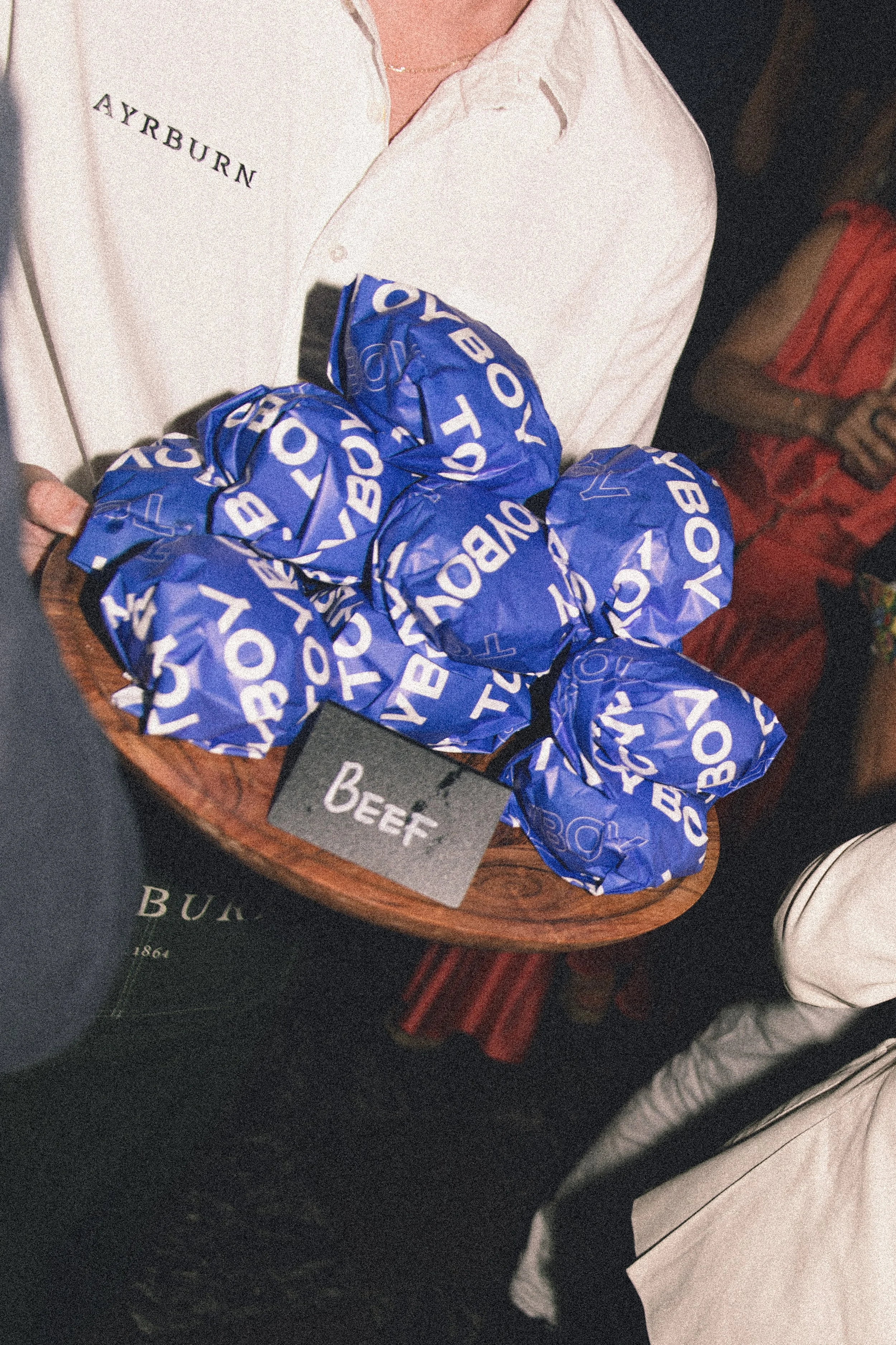 A person holding a tray with several wrapped Burger King Whopper sandwiches in blue paper wrappers. The person is wearing a white shirt with the name 'AYRBURN' embroidered.