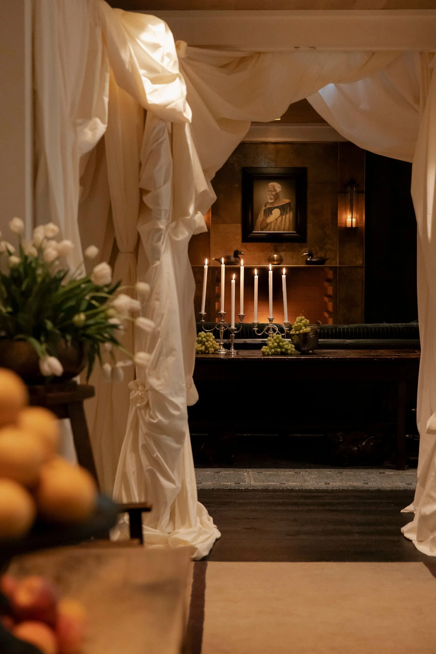 A room decorated with white curtains, floral arrangements, and lit candles on a table. There is a portrait of a distinguished person on the wall behind the table.
