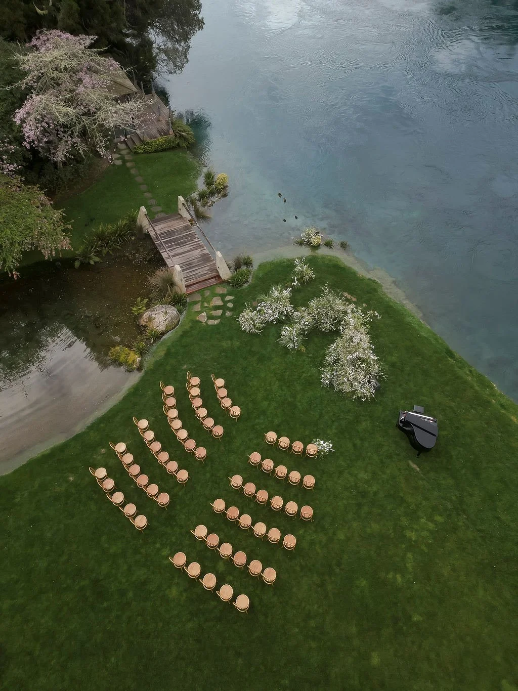 Outdoor lakeside setup with rows of chairs arranged for an event, a grand piano, blooming trees, a wooden dock, and a small pond surrounded by greenery.