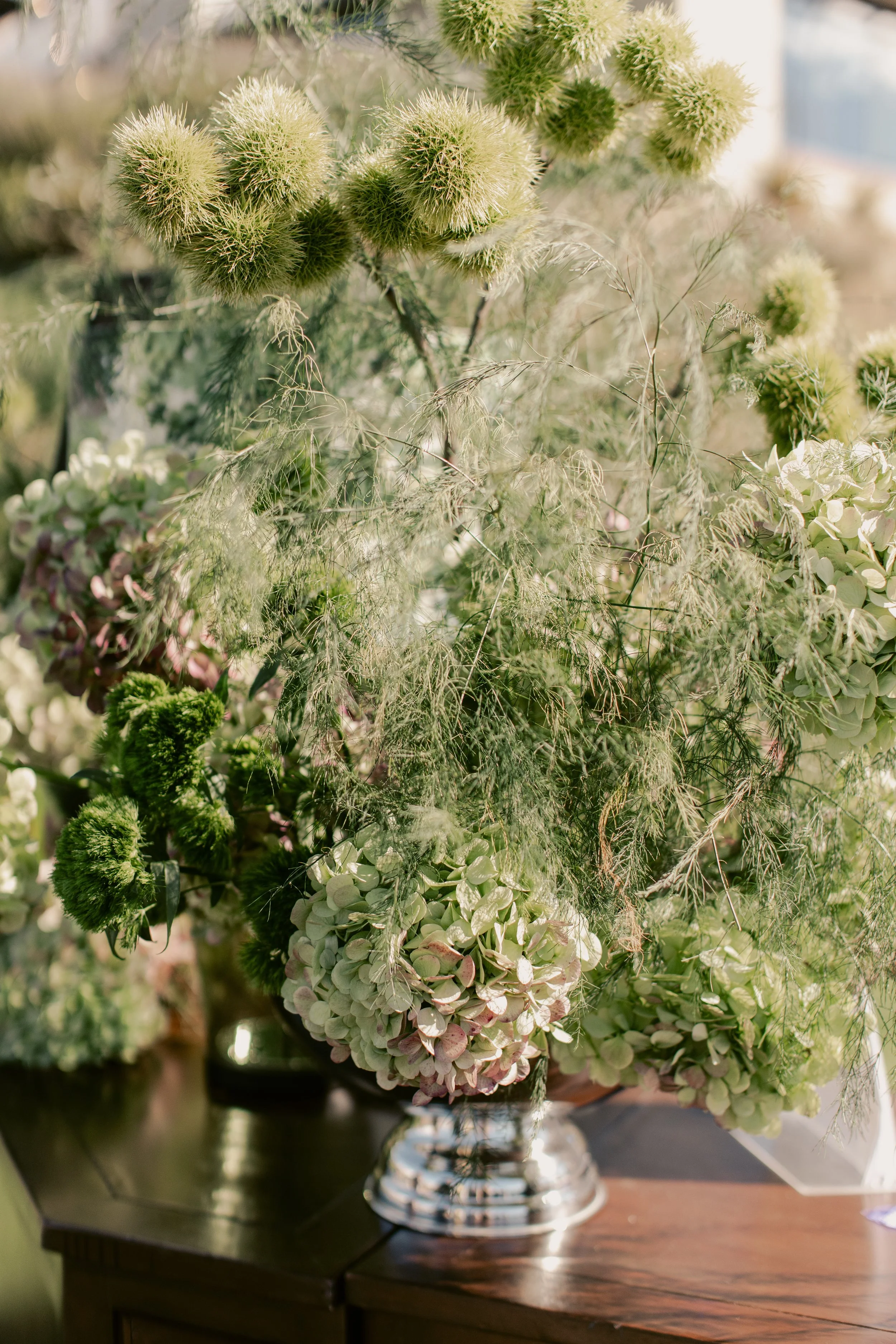 A floral arrangement with pale-green and white flowers, including hydrangeas, in a shiny silver vase on a wooden table.