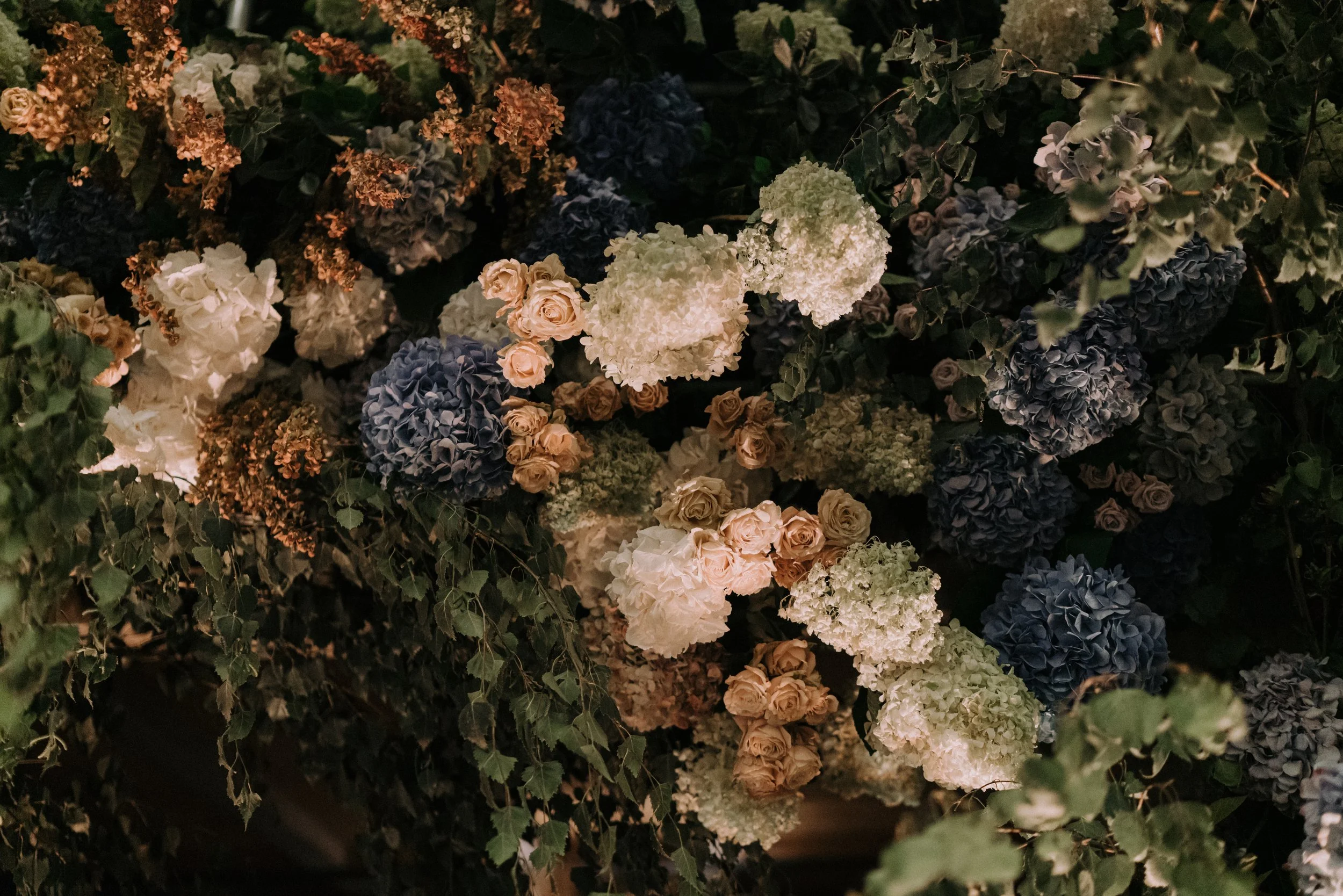 A dense arrangement of various flowers, including hydrangeas and roses, with dark green foliage interwoven among the blooms.