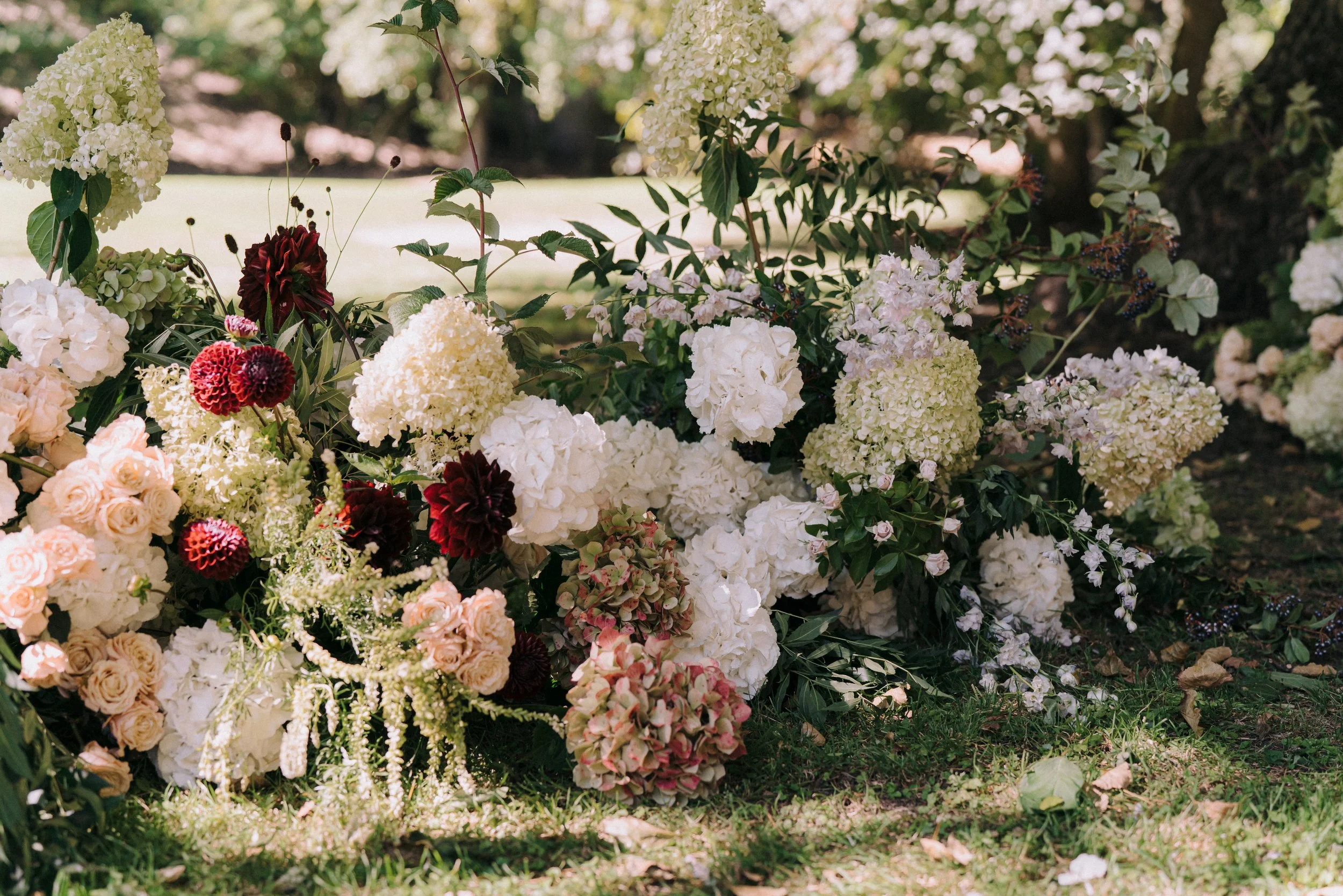 A flower arrangement with white, cream, pink, and burgundy blossoms, including hydrangeas, roses, and other flowers, set outdoors on grass with blurred trees in the background.