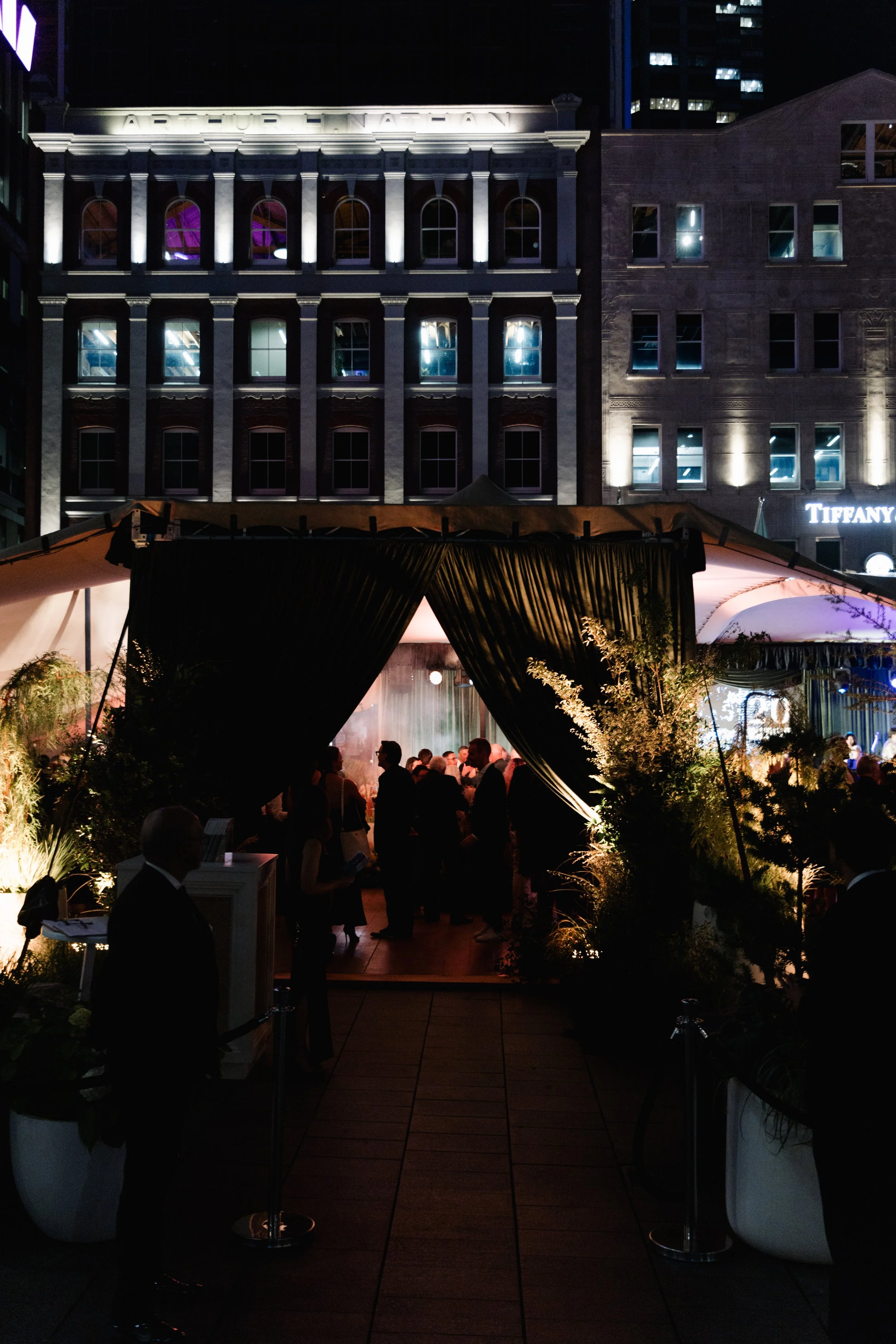 Nighttime scene of a rooftop event with people gathered under a tent. The background features lit-up buildings, including a sign that says 'Tiffany' and a historic-looking building with arched windows and a sign that reads 'Arthur F. Nadeau.'