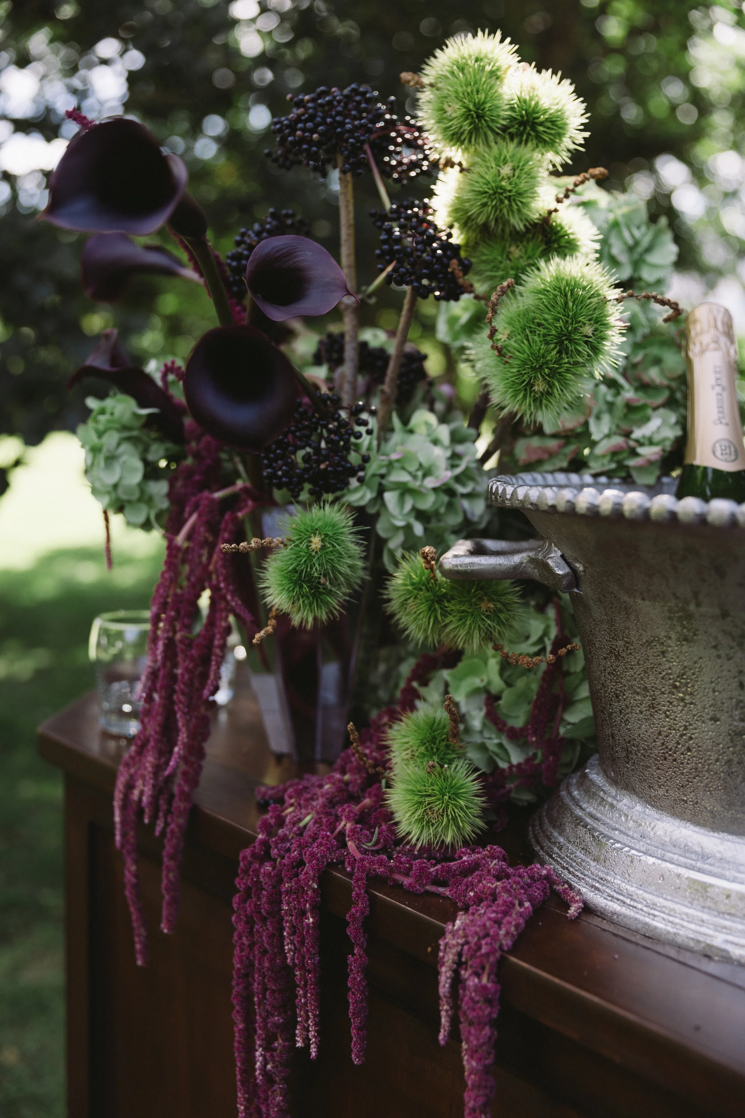 A floral arrangement on a wooden table features green, spiky, round flowers, purple calla lilies, black berries, pinkish-purple amaranthus, and green hydrangeas, set outdoors with blurred greenery and sunlight in the background.