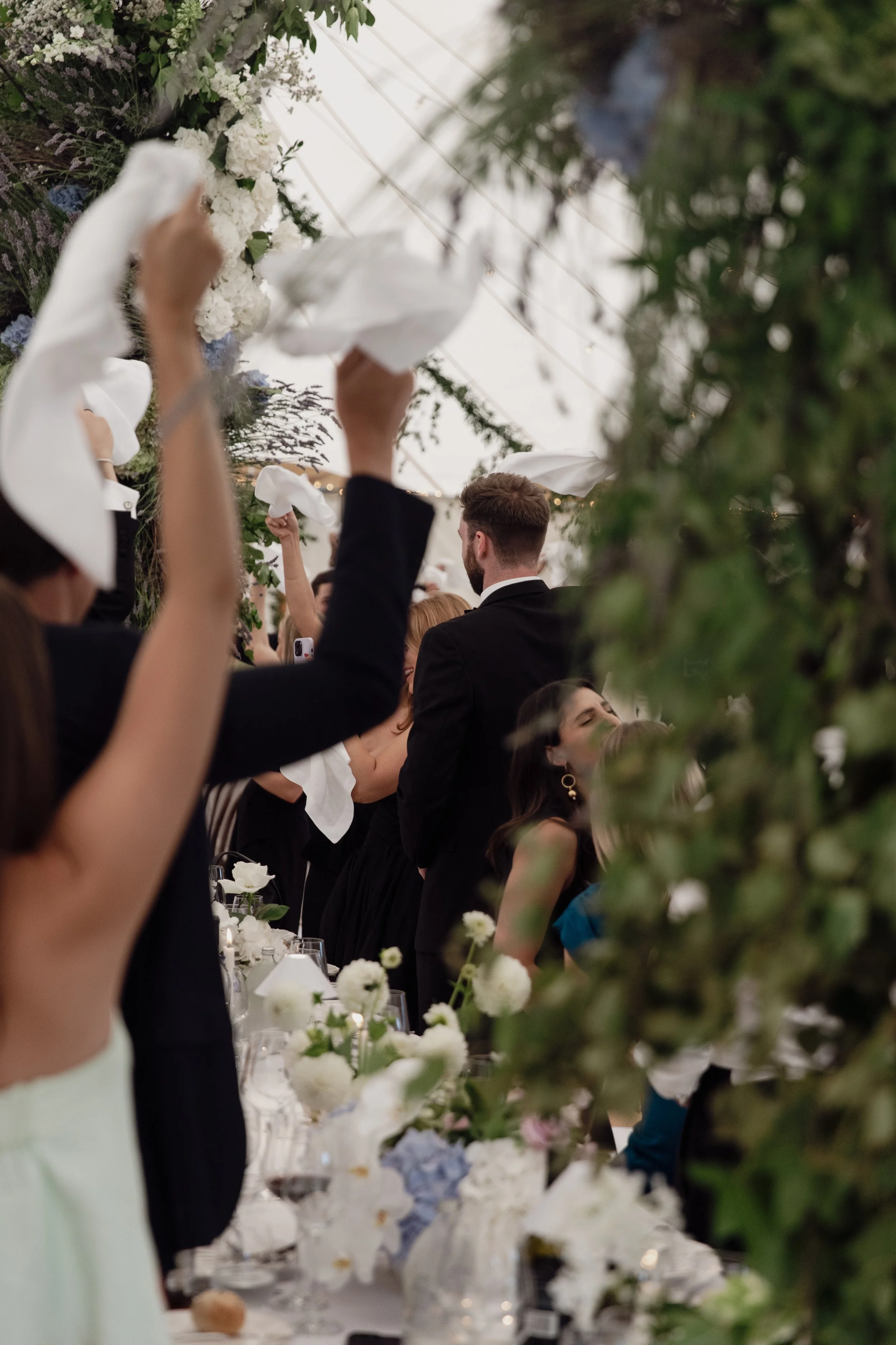 People raising napkins during a formal celebration or wedding reception, decorated with white flowers and greenery.