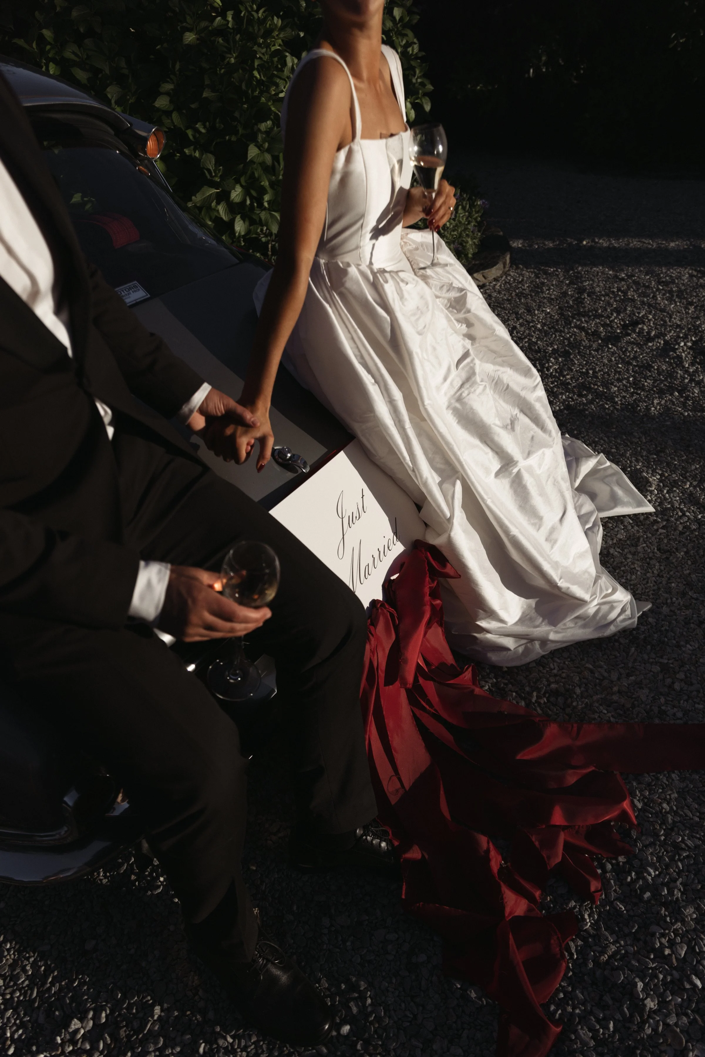 A woman in a white wedding dress holding a glass of champagne sits on a car with a man in a black suit, holding her hand. The scene is outdoors at night with a sign reading 'Just Married' placed on the ground.