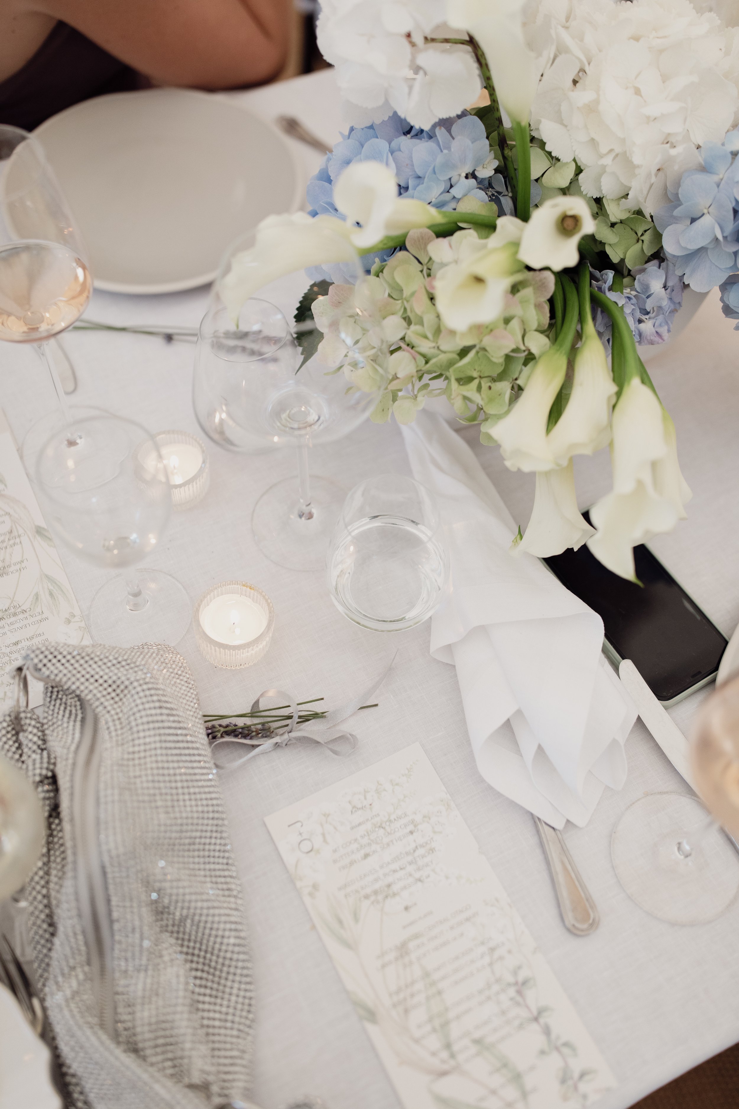 A centerpiece with white and light blue hydrangeas and white calla lilies on a table set for an event, with candles, glasses, a menu, and a checkered cloth.