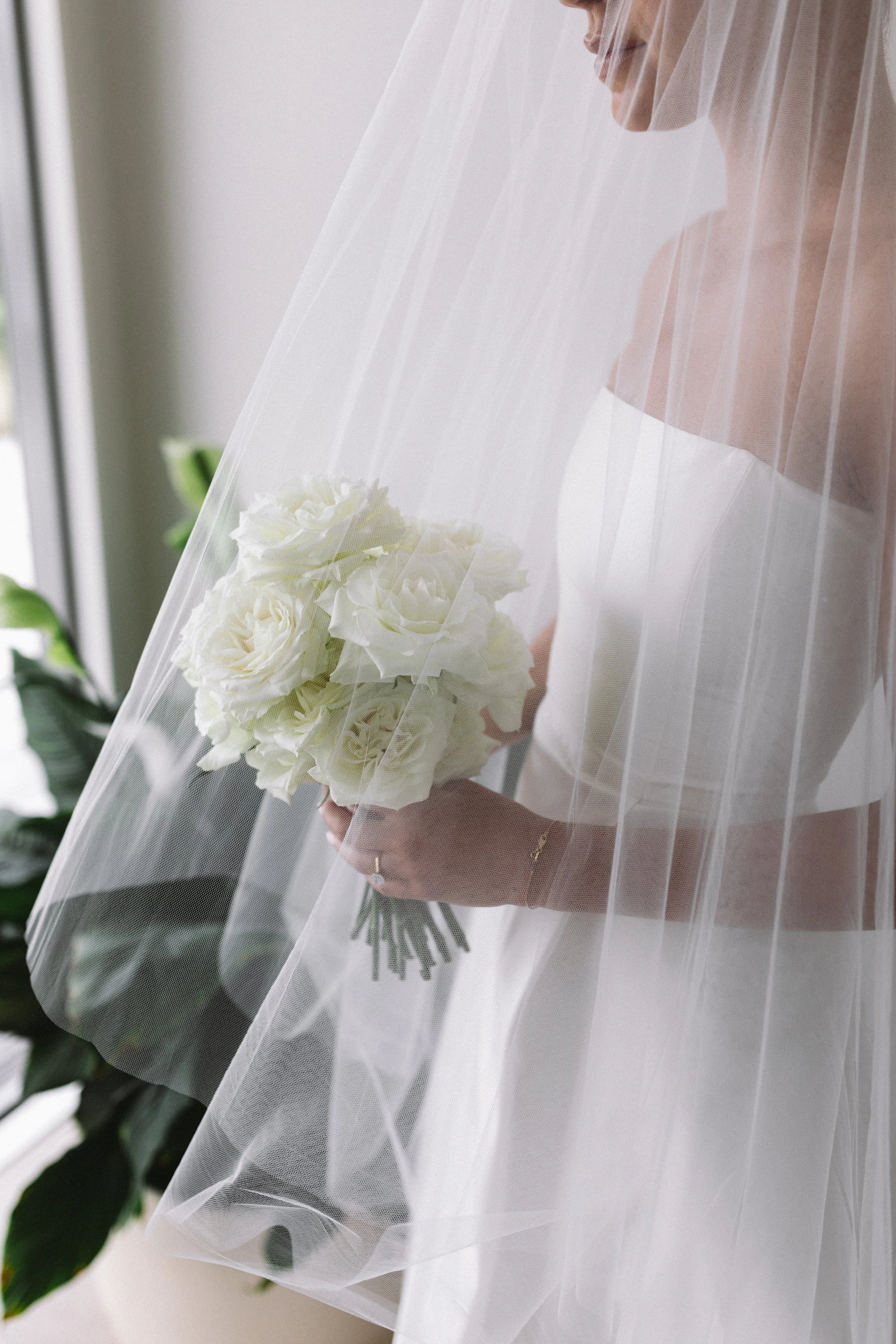 A bride holding a bouquet of white roses, wearing a wedding dress and a veil, with a blurred background of green plants.