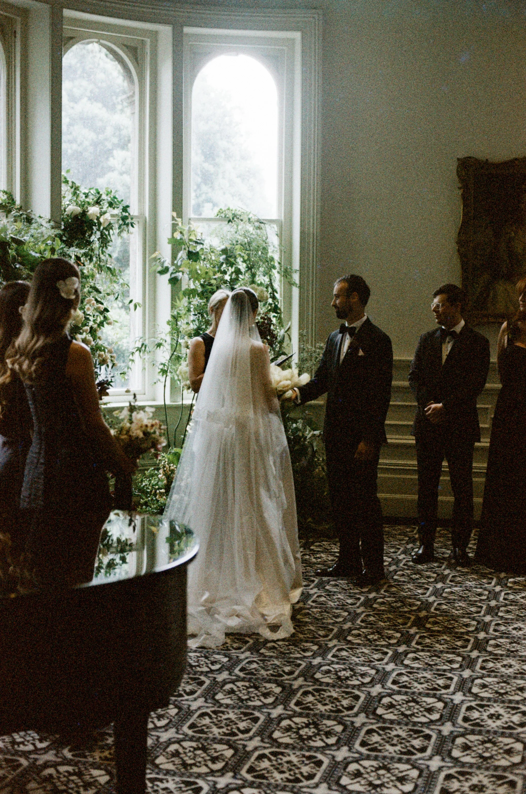 A wedding ceremony with the bride and groom standing together, exchanging rings in a room with large windows, decorated with flowers and plants, and patterned carpet.