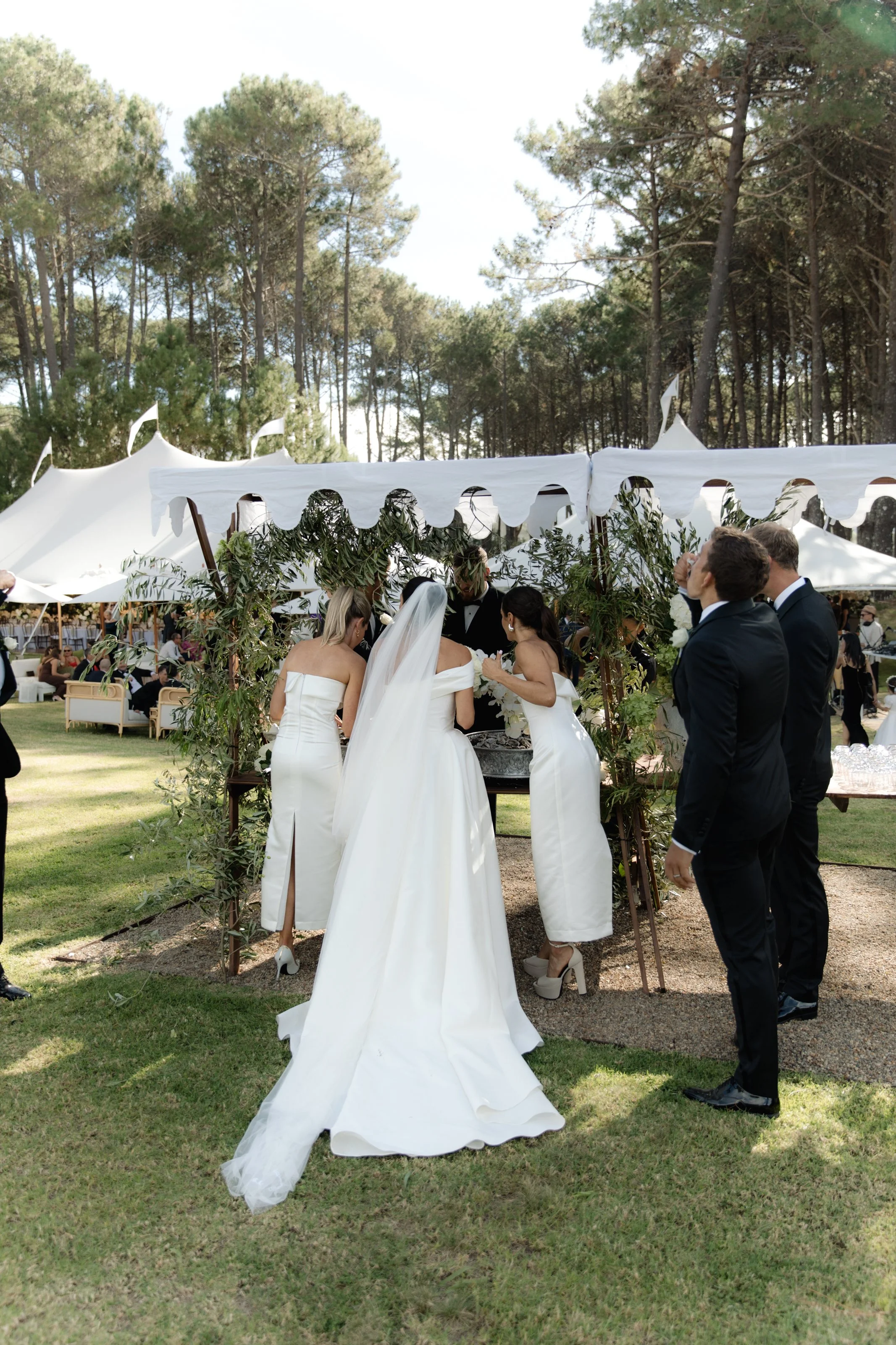 Wedding ceremony outdoors with a bride and groom standing under a white canopy decorated with greenery, surrounded by guests and forest in the background.