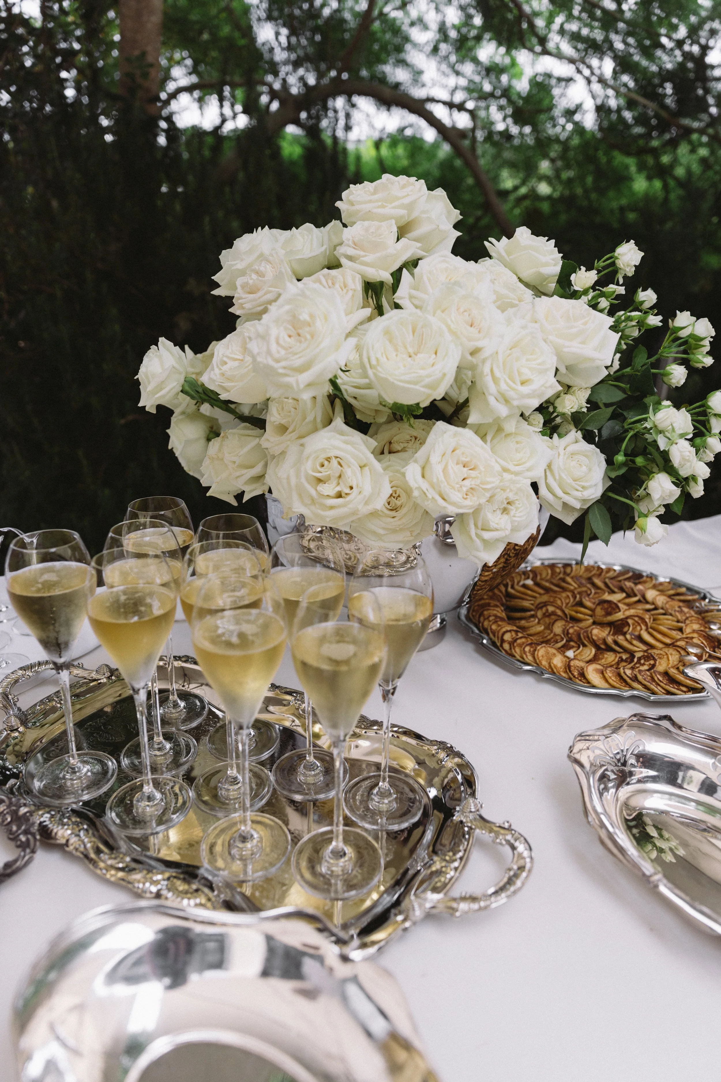 A large bouquet of white roses and smaller white flowers on a table with silver trays of champagne and snacks, set outdoors with green foliage in the background.