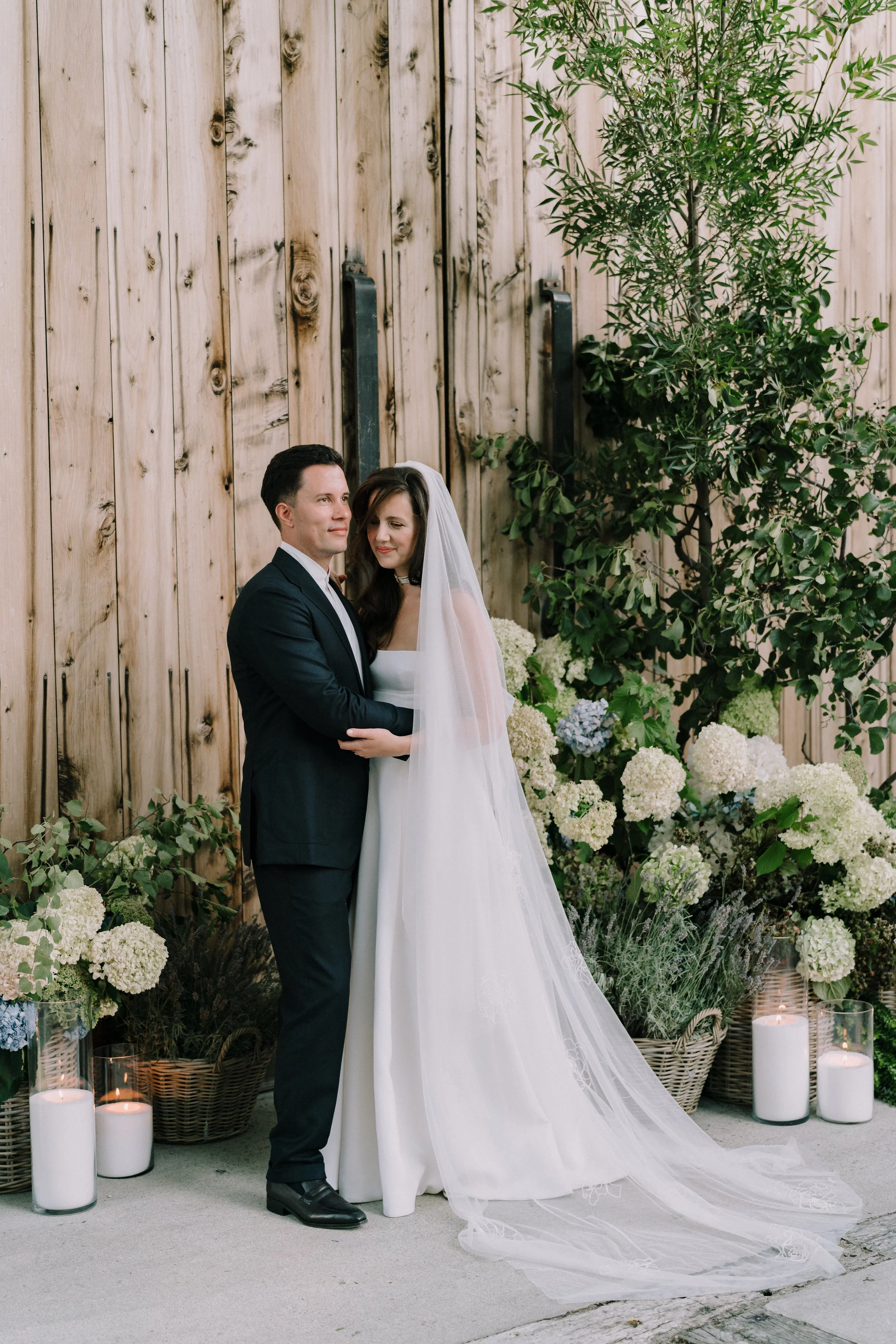 A newlywed couple stands close together, the groom in a black suit and the bride in a white wedding gown with veil. They are surrounded by white and green floral arrangements and candles on the floor, with a wooden wall and lush green trees in the background.