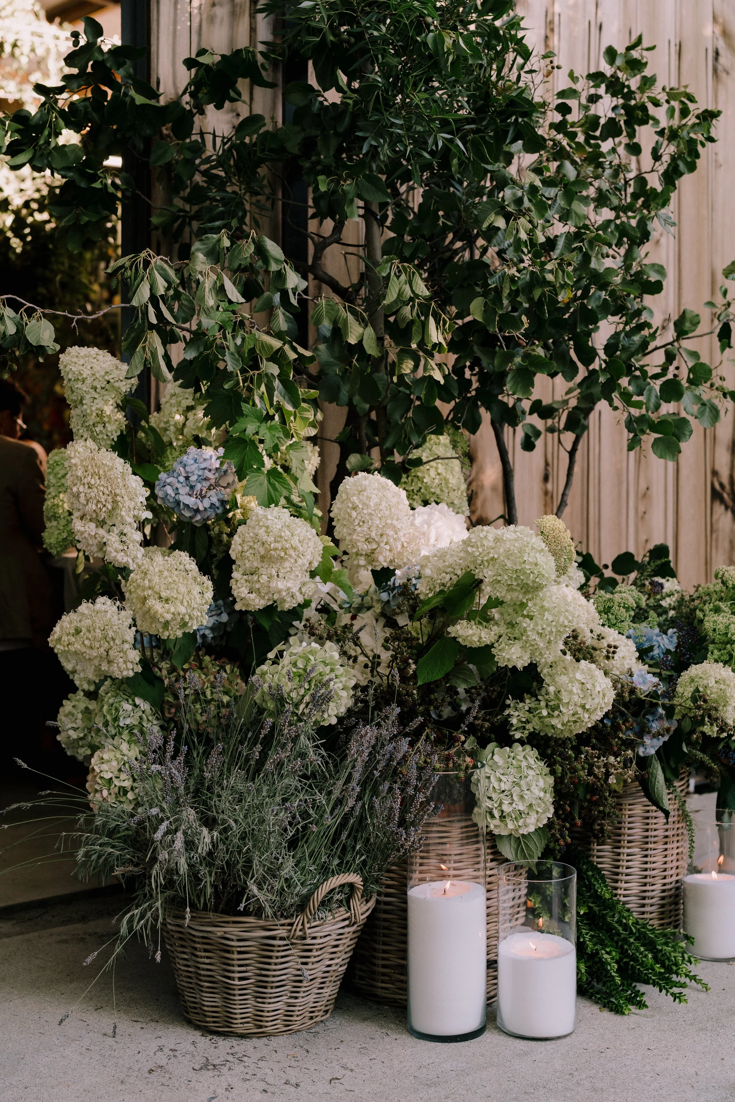 Decorative floral arrangement with white hydrangeas, greenery, and lavender in rattan baskets, accented with white candles in glass holders, against a wooden fence background.