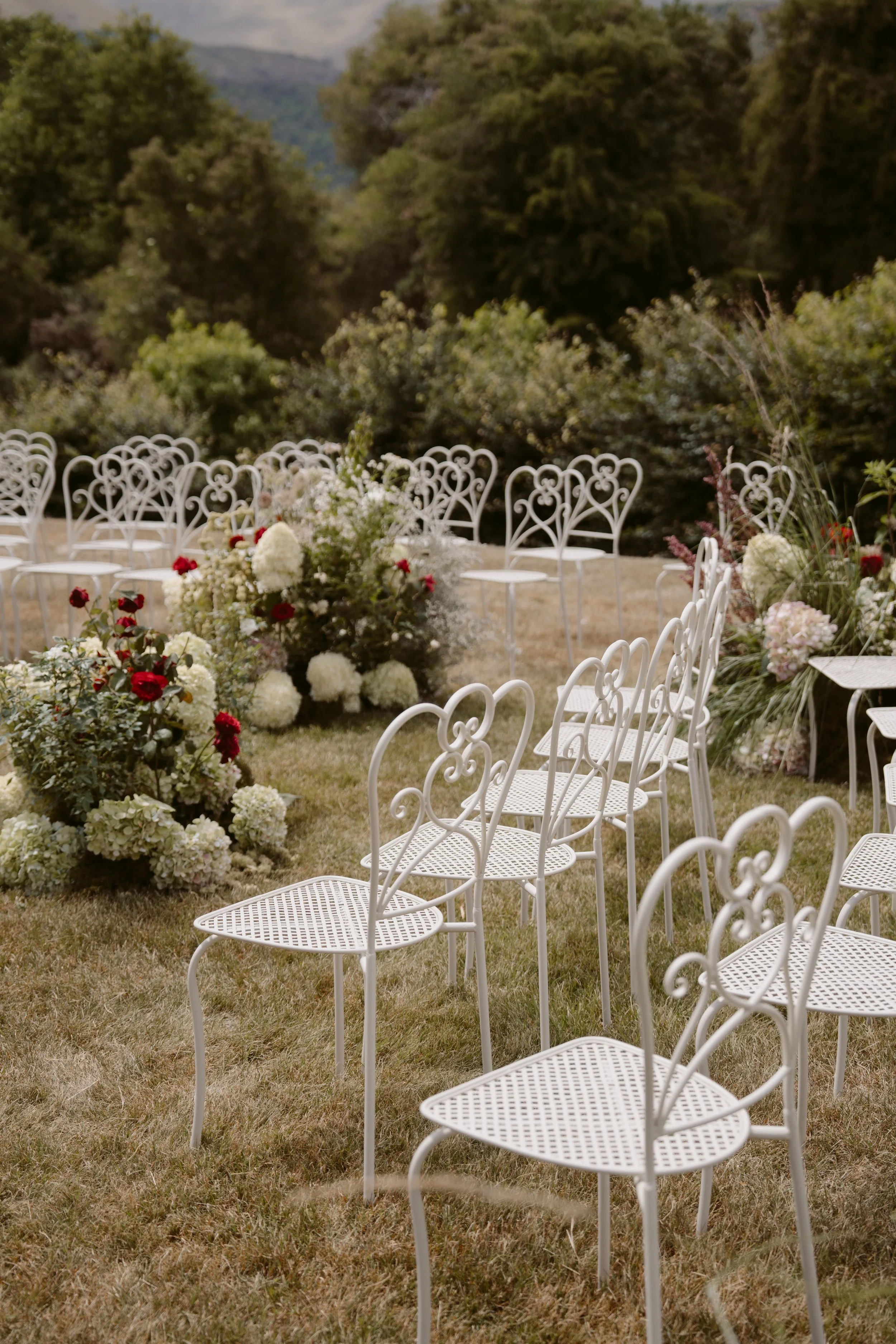 Outdoor wedding ceremony setup with white ornate chairs and floral arrangements on a grassy area, surrounded by trees and mountains.