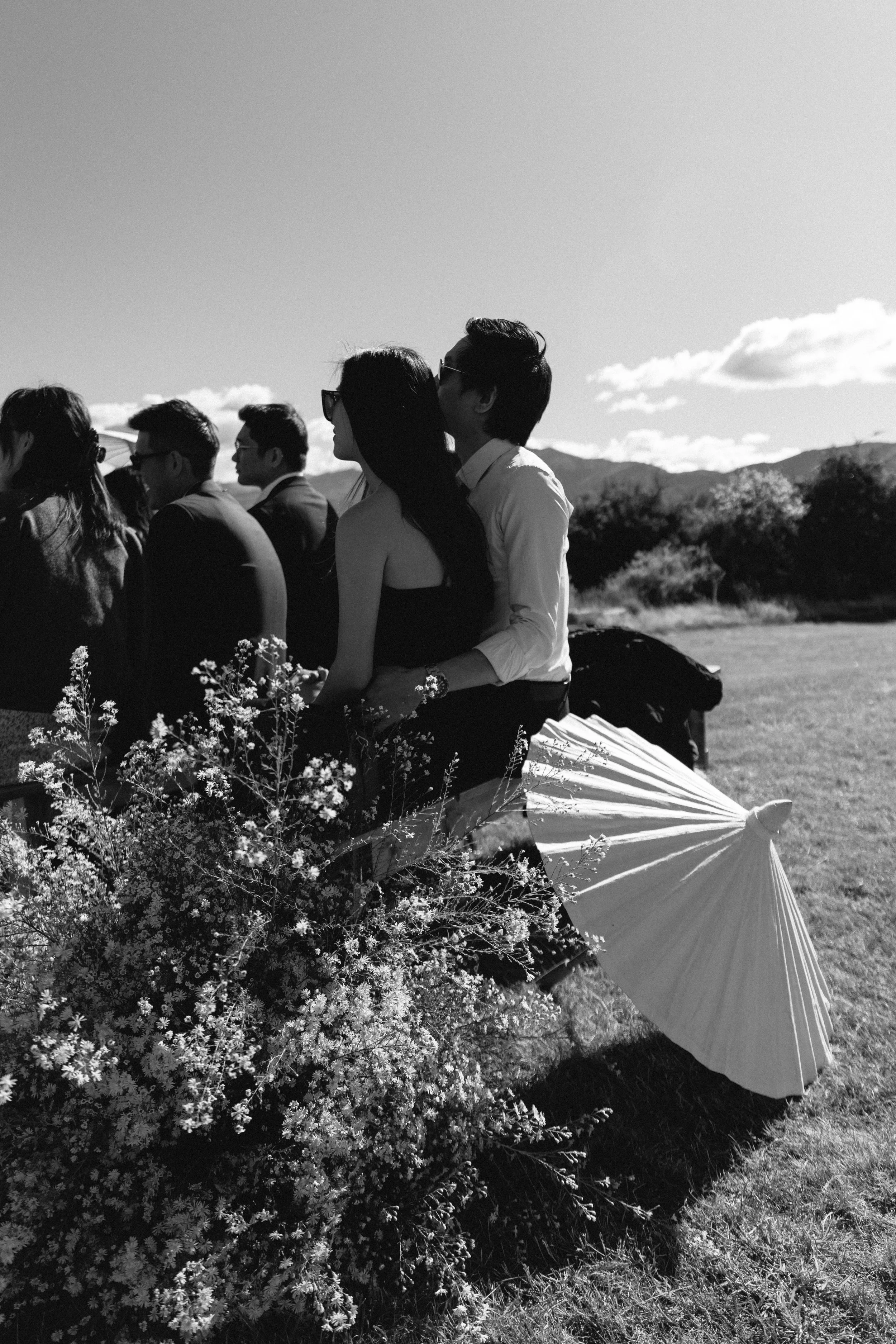 A group of people in formal attire, sitting outdoors in a scenic landscape with mountains and trees, under a clear sky. There is a white umbrella and flowers in the foreground.
