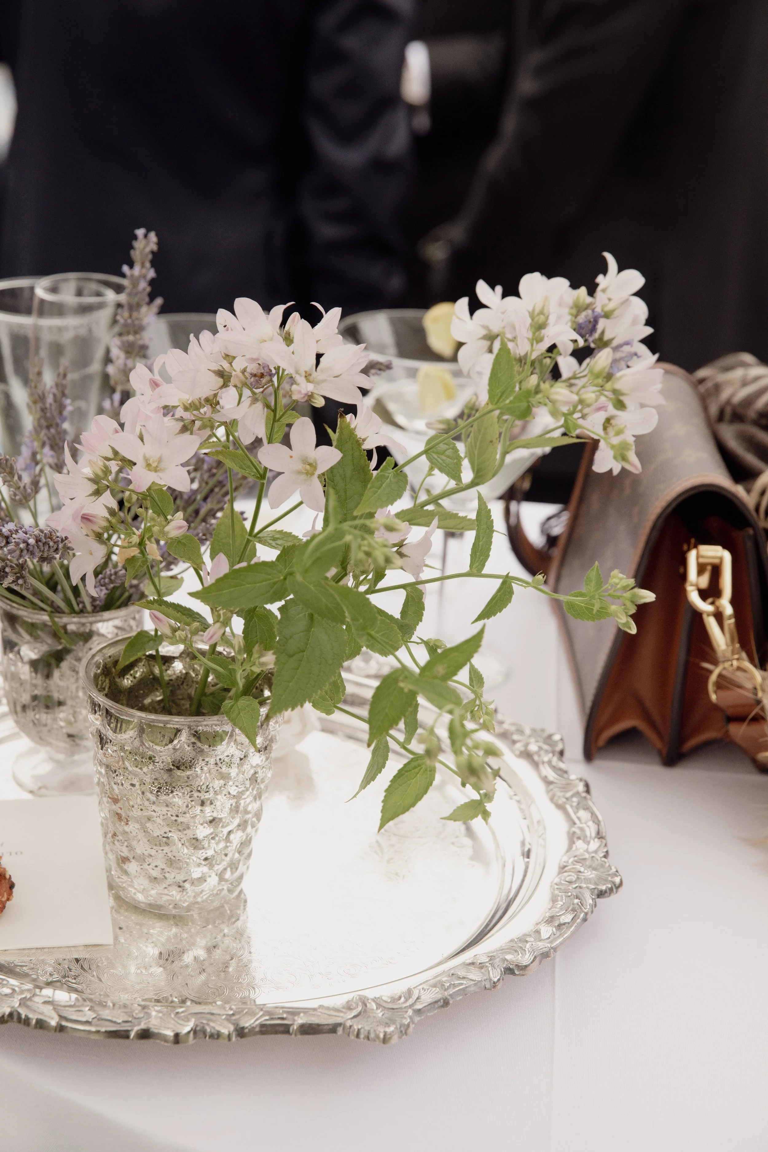 Pink and white flowers in a textured glass vase on a silver tray with a lace edge, set on a white tablecloth, with a blurred background of glassware, a brown bag, and a dark backdrop.