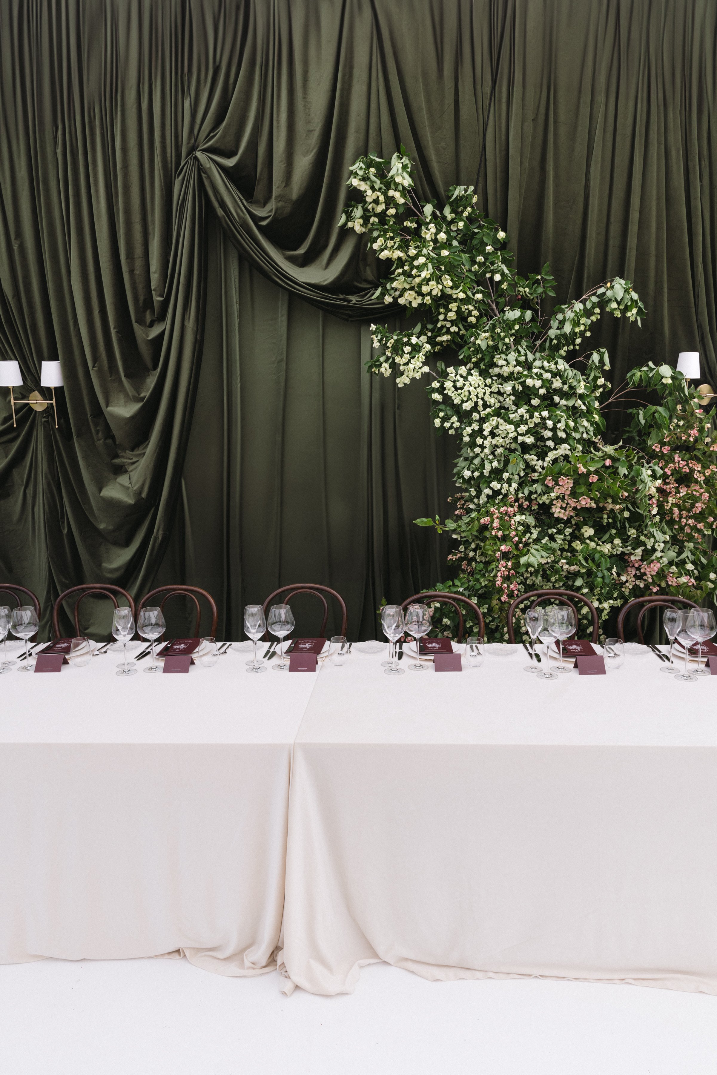 A large banquet table with a white tablecloth, set with wine glasses and menus, in front of a backdrop of dark green curtains and a tall arrangement of white and pink flowers.