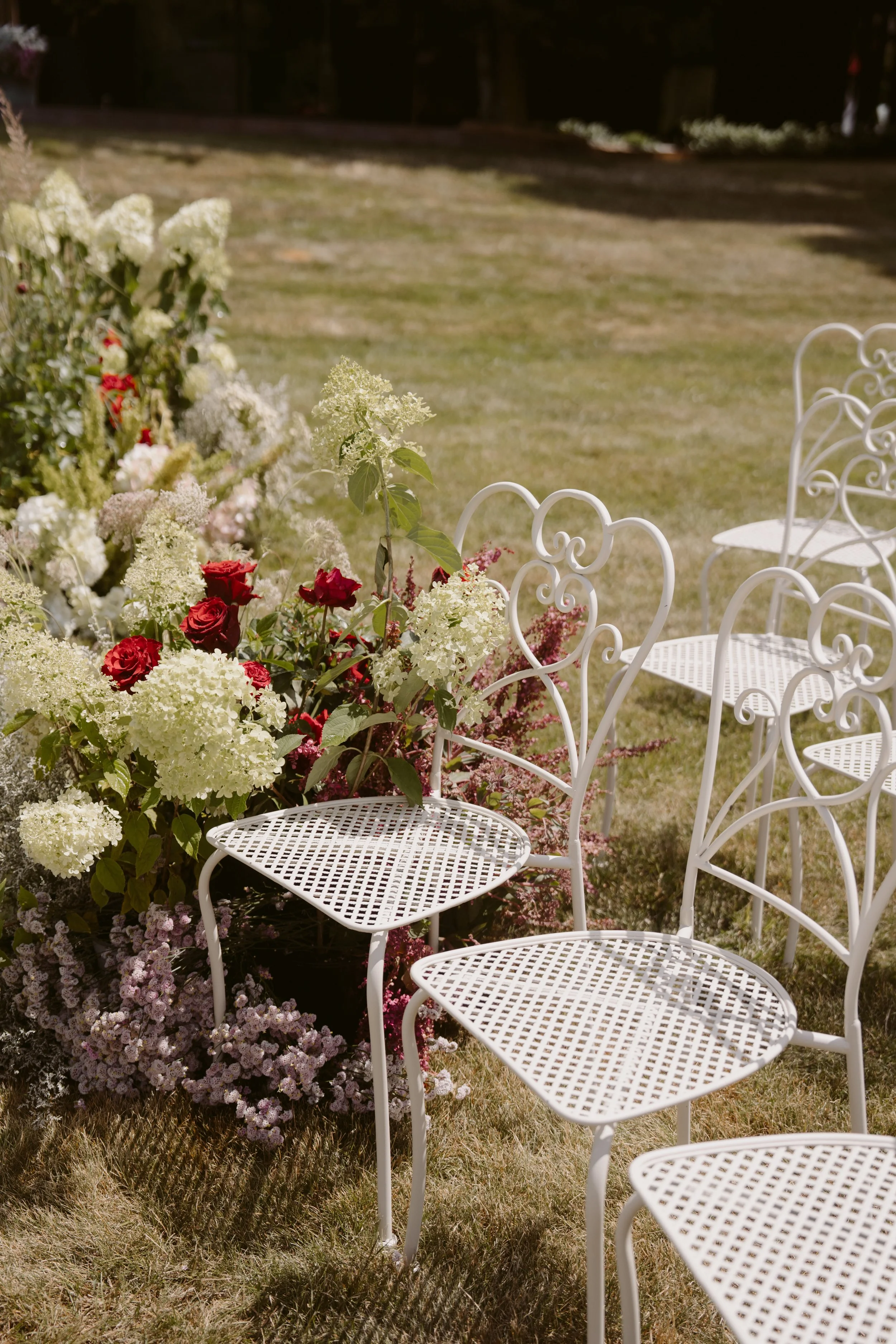 White wrought iron chairs arranged outdoors on a grassy lawn, near a flower bed with white, red, pink, and purple flowers.