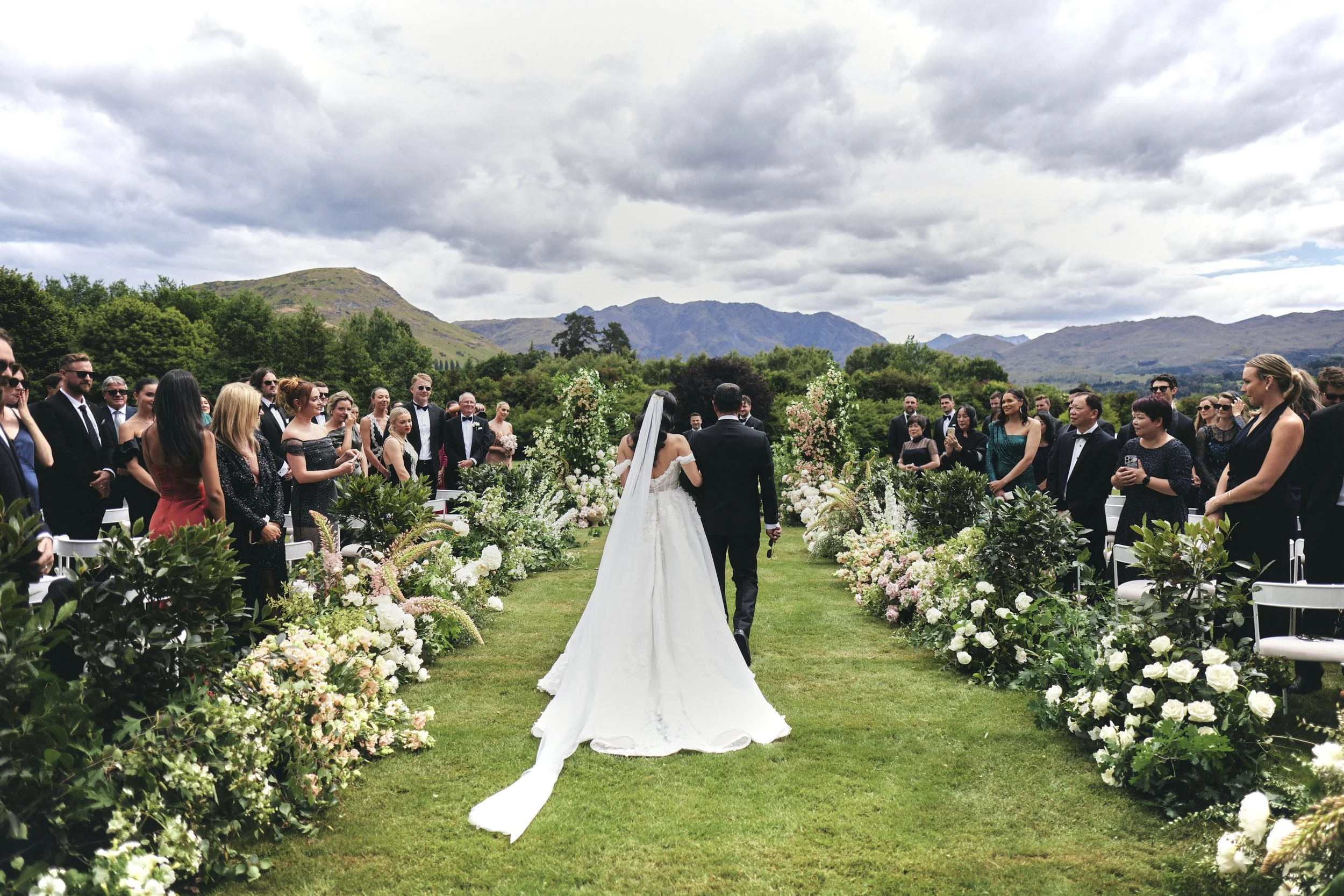 Outdoor wedding ceremony with bride and groom facing each other, surrounded by guests and floral arrangements, mountain scenery in the background under cloudy sky.