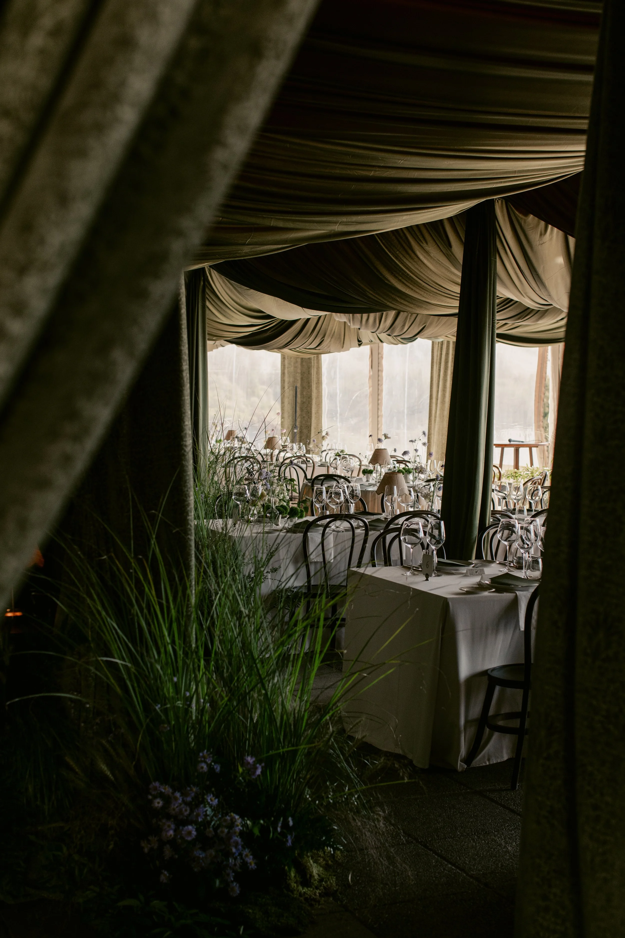 A banquet setup inside a venue with draped ceiling, tables with white tablecloths, wine glasses, and floral centerpieces, viewed through some curtains and greenery.