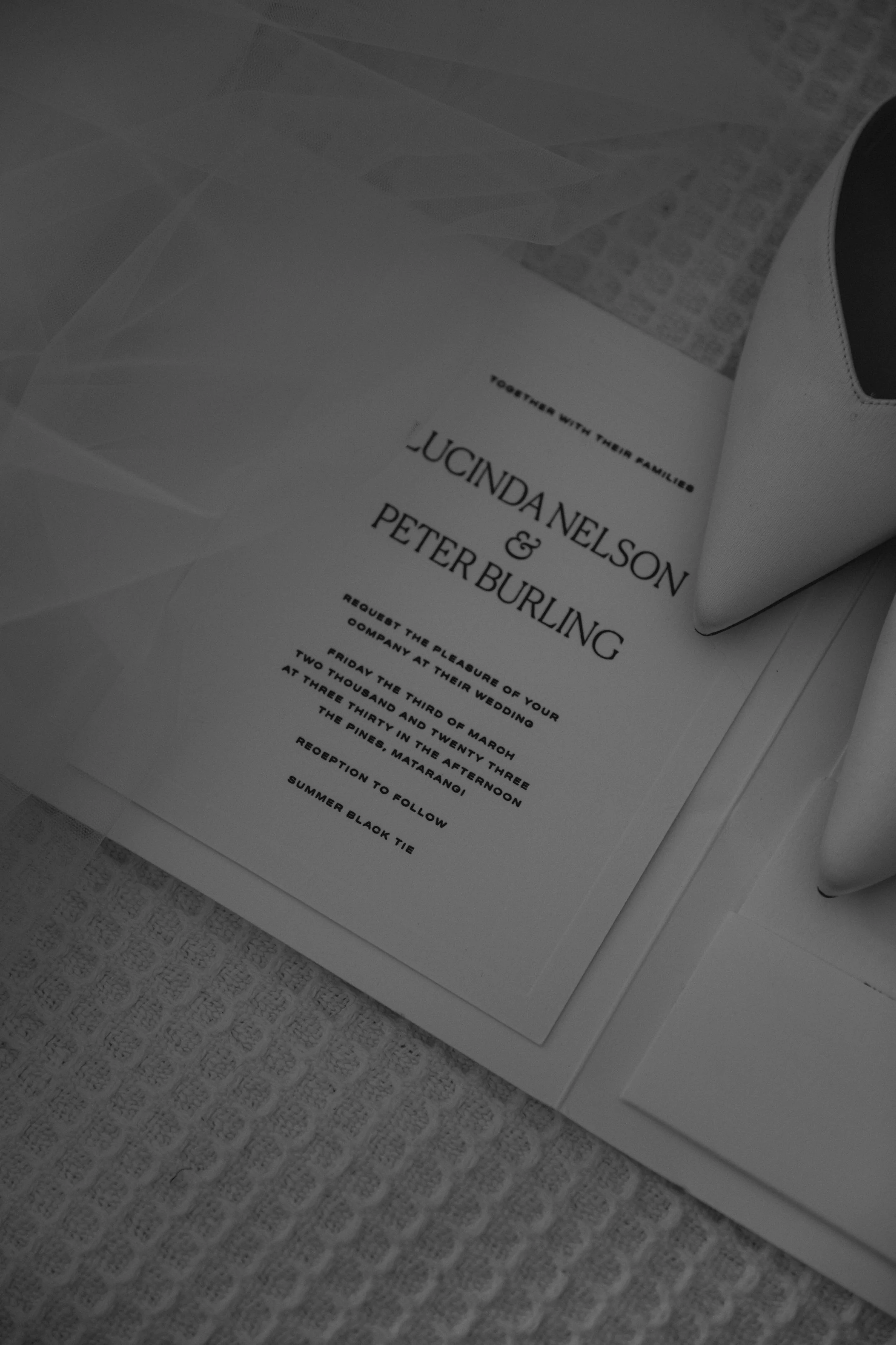 Close-up of an invitation or program card for Lucinda Nelson and Peter Burling's wedding, placed on a textured surface with a white shoe partially visible on the right side.