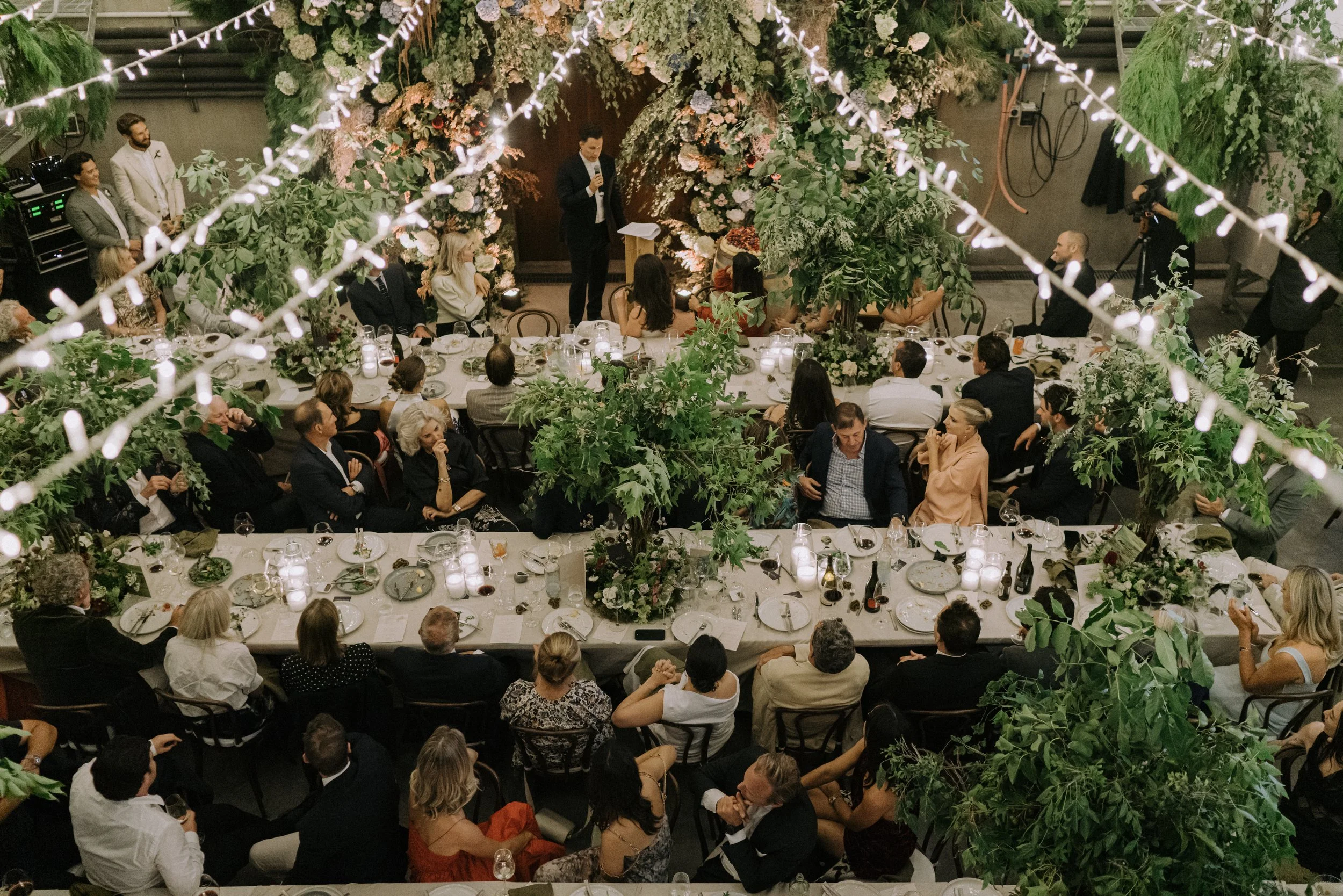 A formal event or wedding reception with guests seated around a long decorated table filled with plates, glasses, and candles. The area is adorned with lush green foliage and large floral arrangements, and string lights hang overhead. A man in a dark suit is speaking at a podium decorated with flowers, with other individuals standing and sitting nearby.