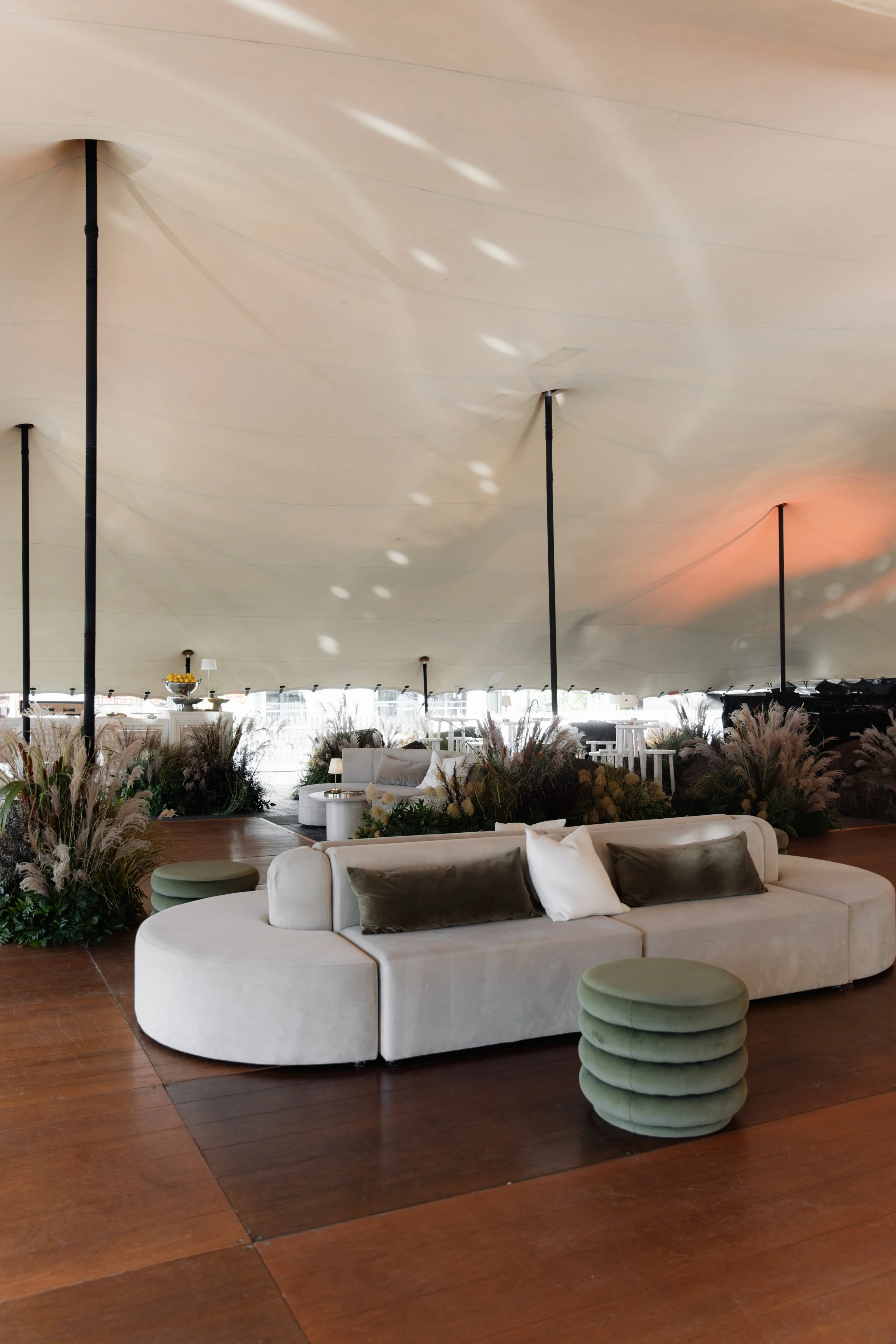 A spacious indoor lounge with modern white sofas, green ottomans, and an abundance of plants and floral arrangements, all under a large, curving ceiling with soft lighting.