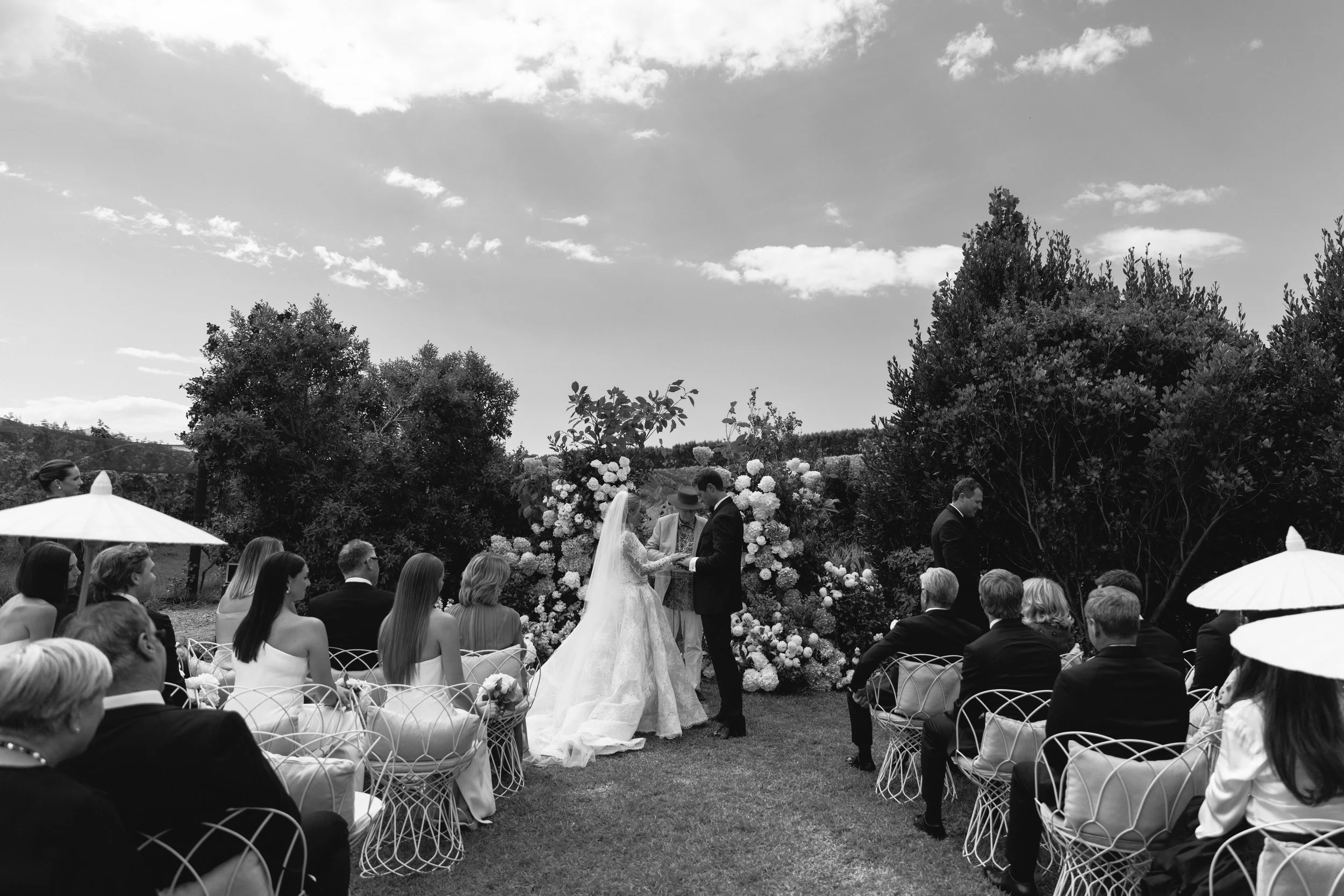 A black and white photo of a wedding ceremony outdoors with guests seated on chairs, the bride and groom exchanging vows, surrounded by flowers and trees under a partly cloudy sky.