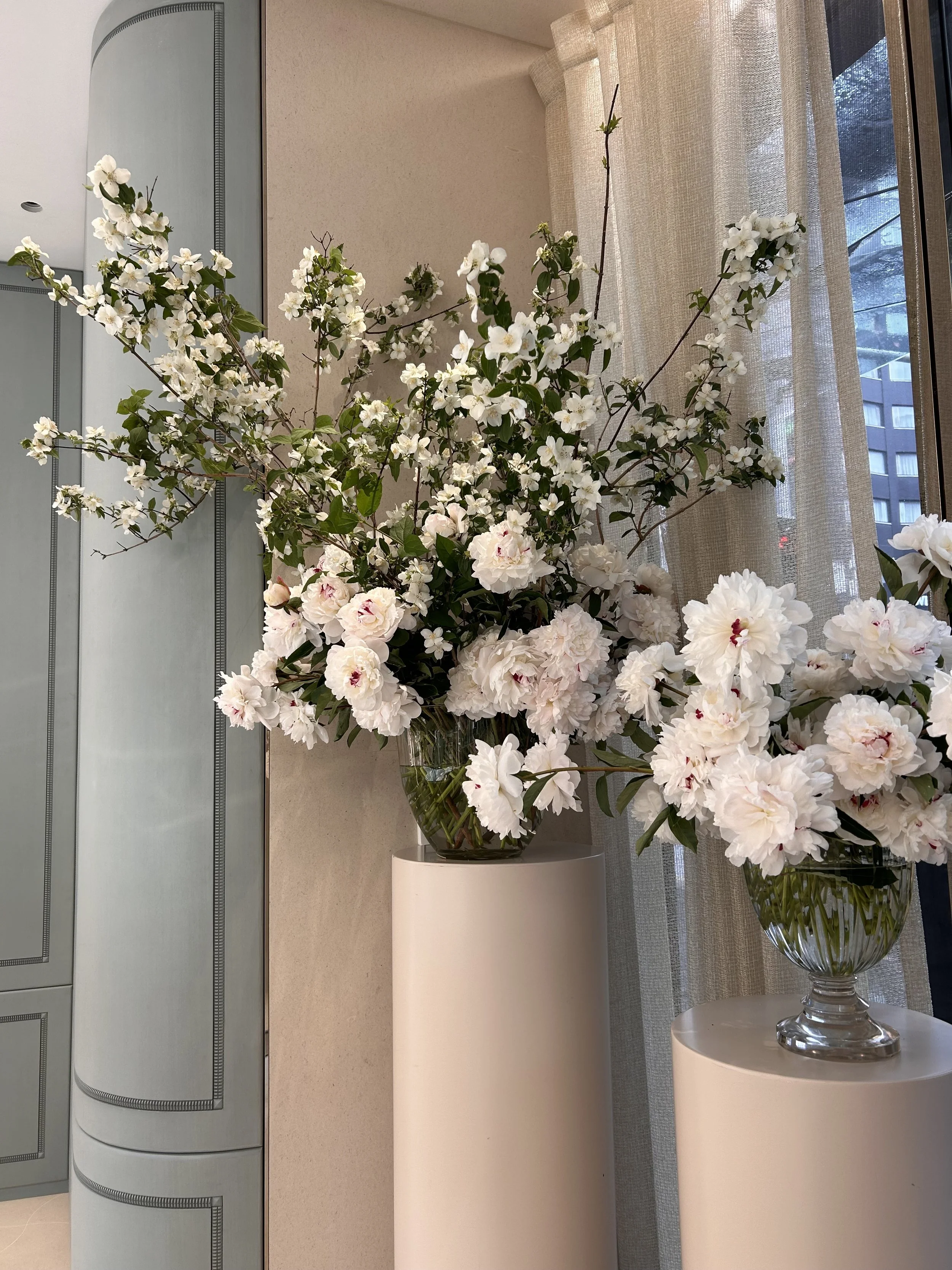 Two glass vases filled with white flowers, placed on white cylindrical pedestals in front of a background with a window and beige curtains.