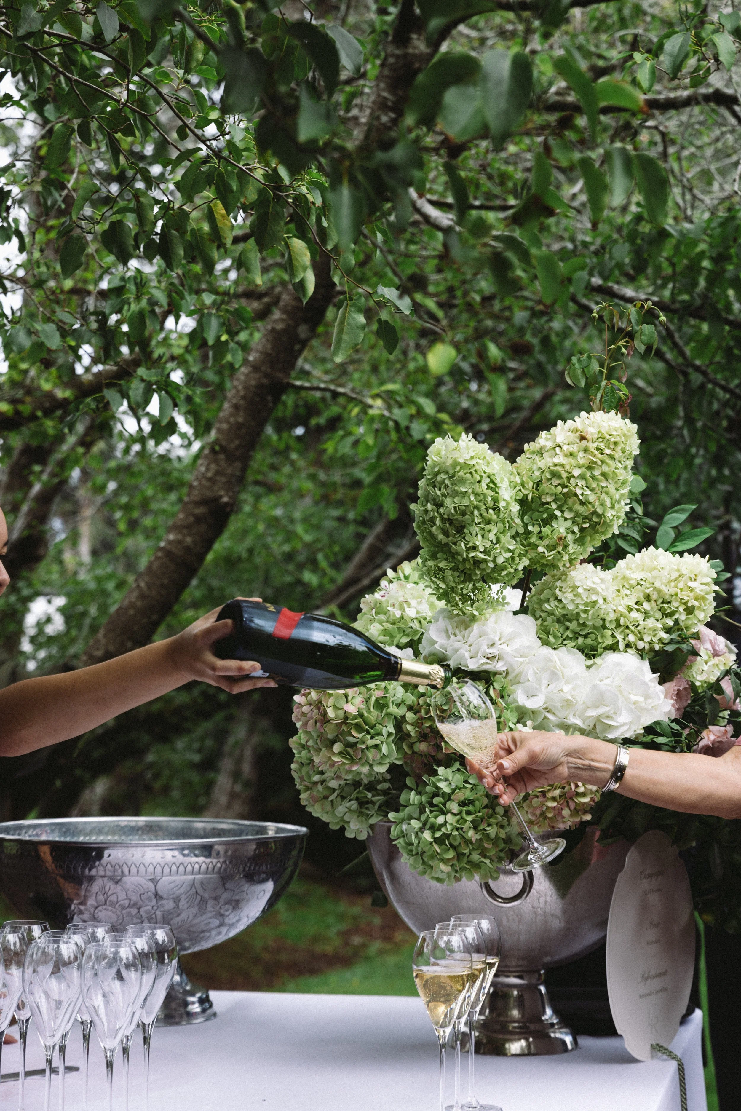 People pouring champagne at a celebration with floral arrangements and champagne glasses on a table outdoors.