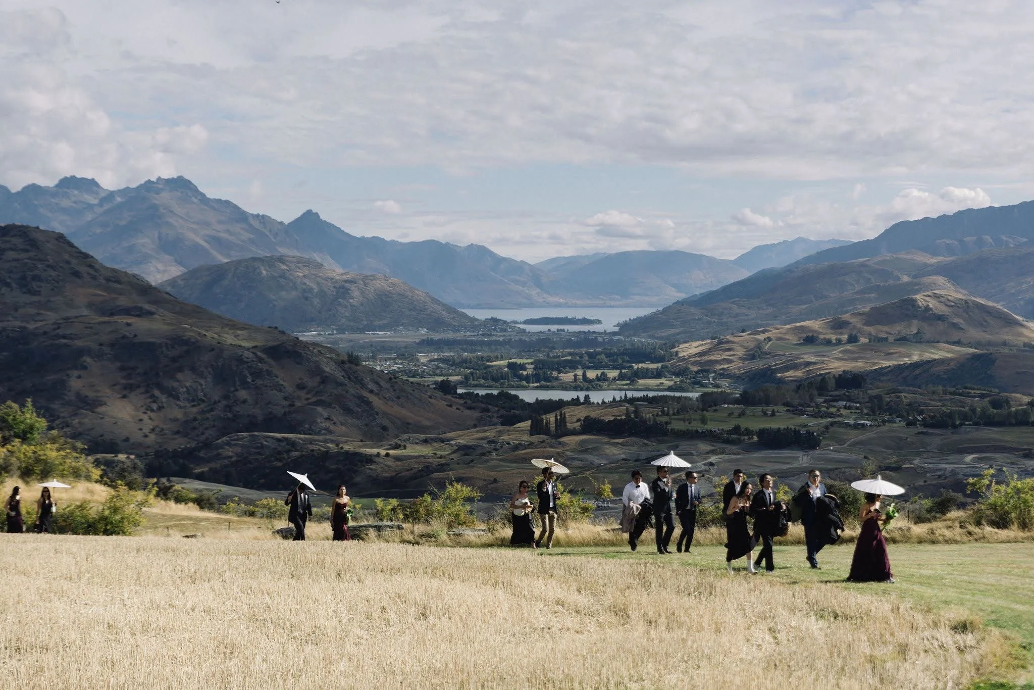 A group of people dressed formally with umbrellas walking through a grassy field, with mountains, a lake, and cloudy sky in the background.