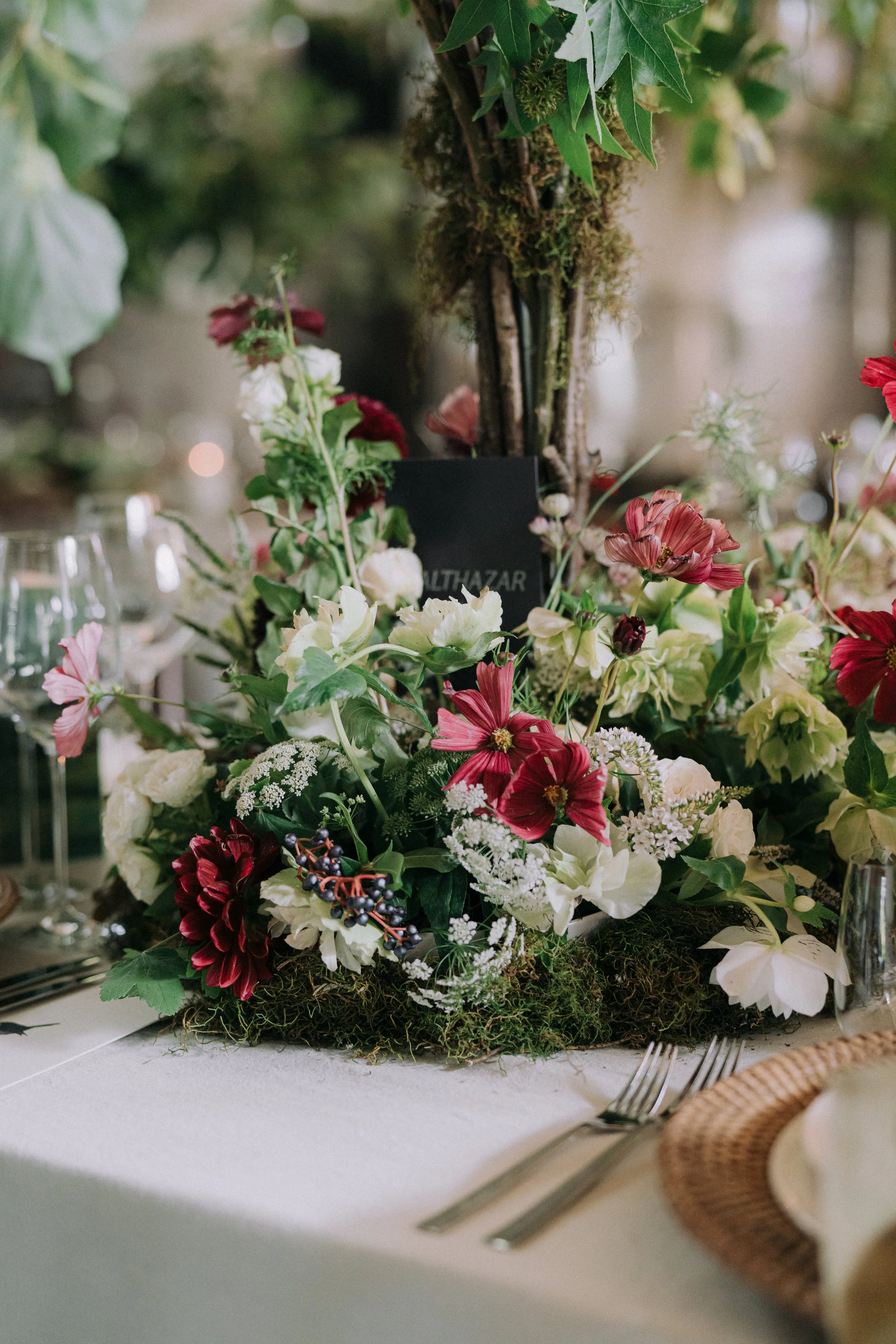A floral centerpiece on a dining table with white and red flowers, greenery, and moss, surrounded by tableware including forks and a woven placemat.