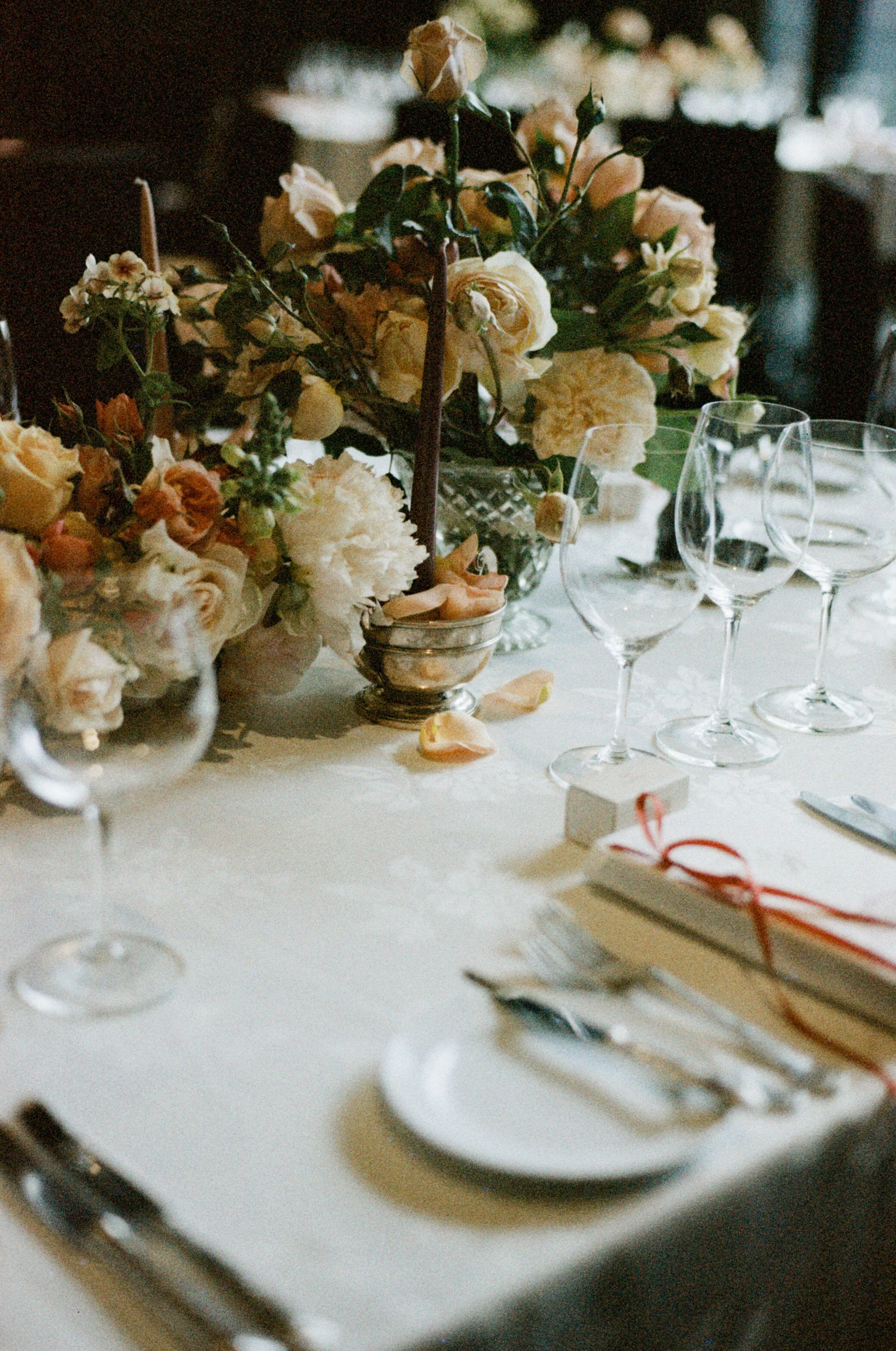A table set for a formal event with a centerpiece of cream and peach flowers, multiple empty wine glasses, and tableware, including a menu or note tied with a red ribbon.