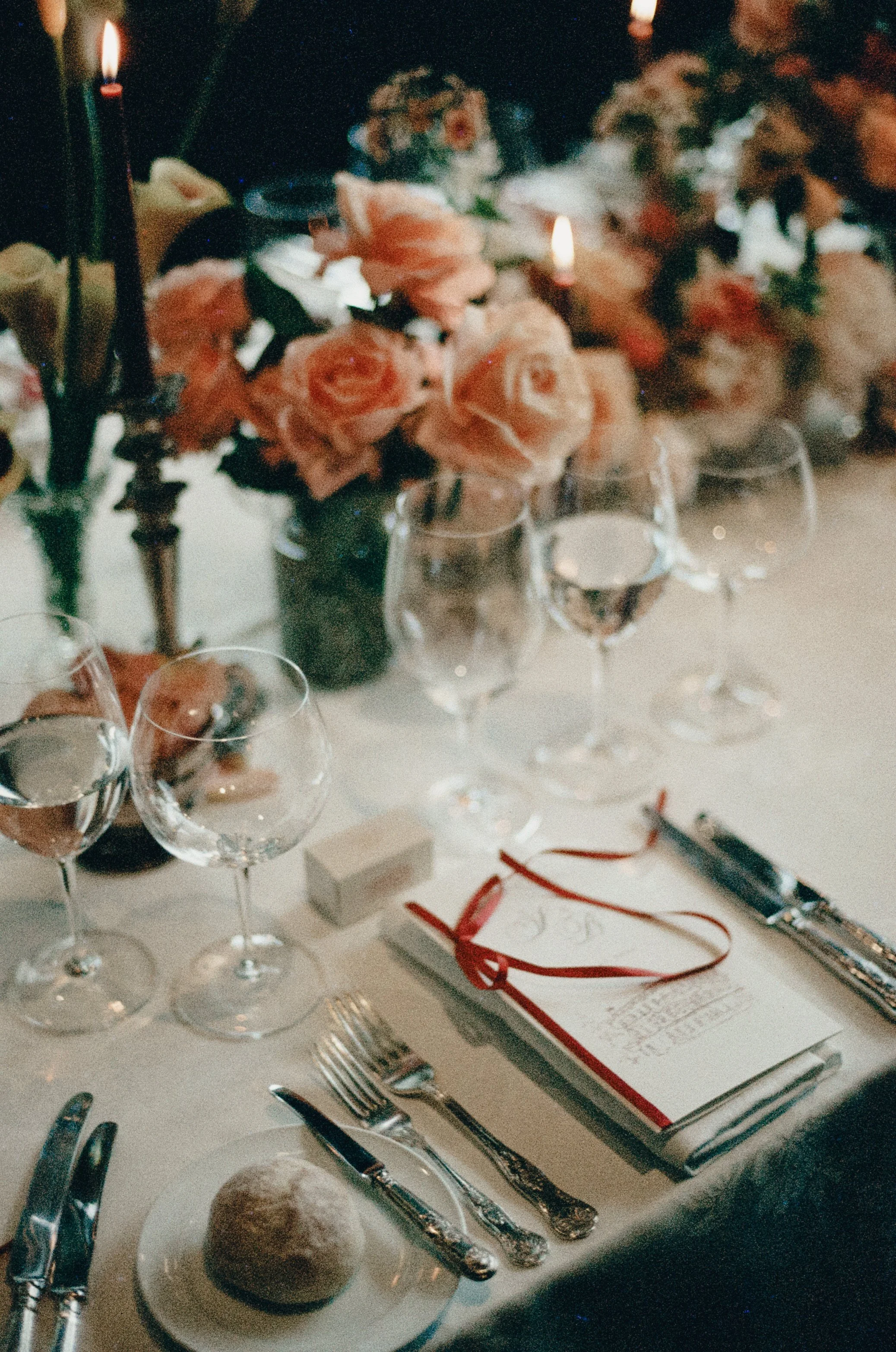 A formal dining table setup with multiple wine glasses, silverware, a bread roll on a plate, and a floral centerpiece. There is a white menu with red accents, and a pair of red glasses on the table, indicating a celebratory or special event.