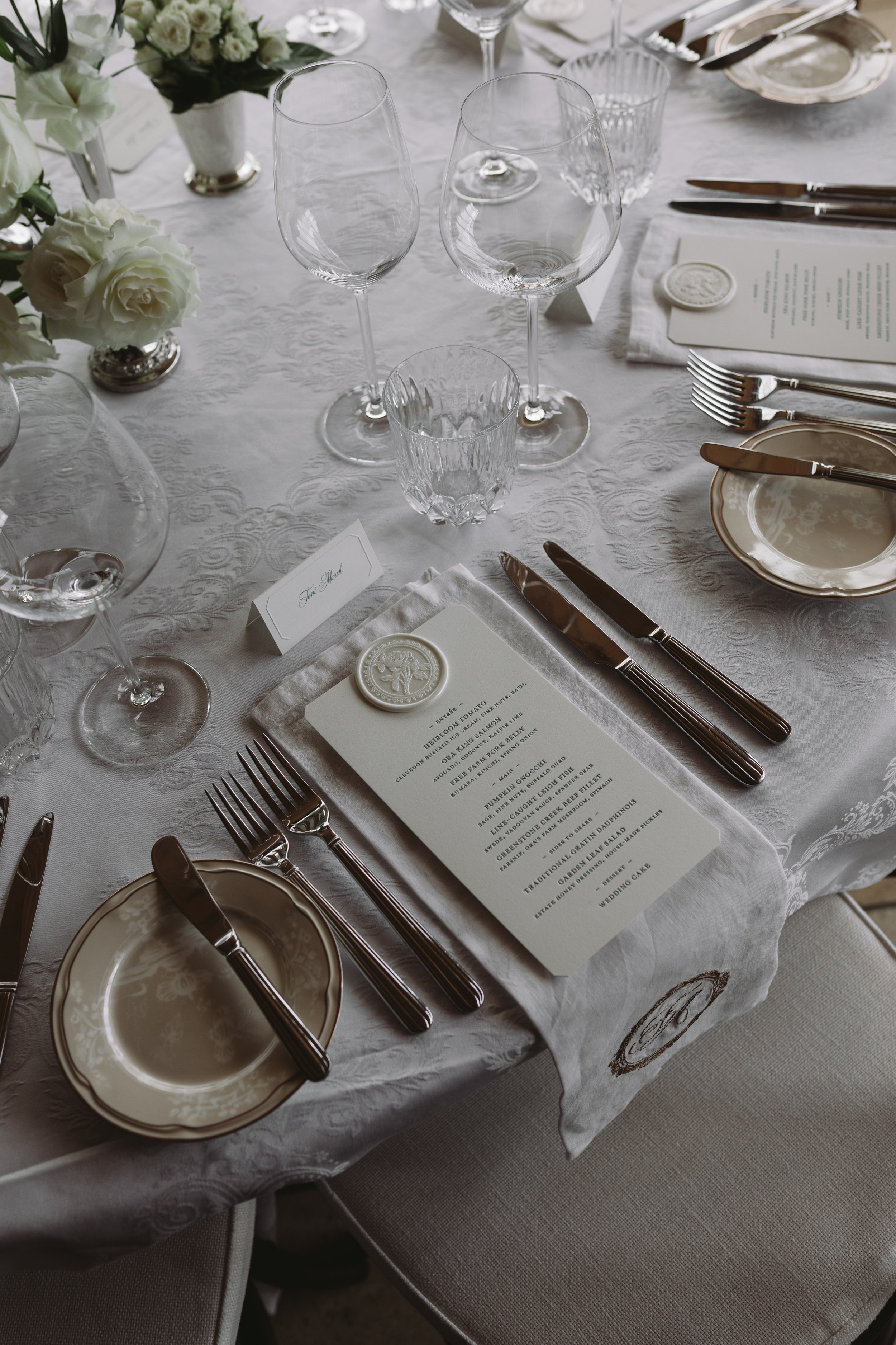 A formal dining table set with white tablecloth, empty wine glasses, silverware, a menu, floral centerpieces, and place cards for an elegant event.
