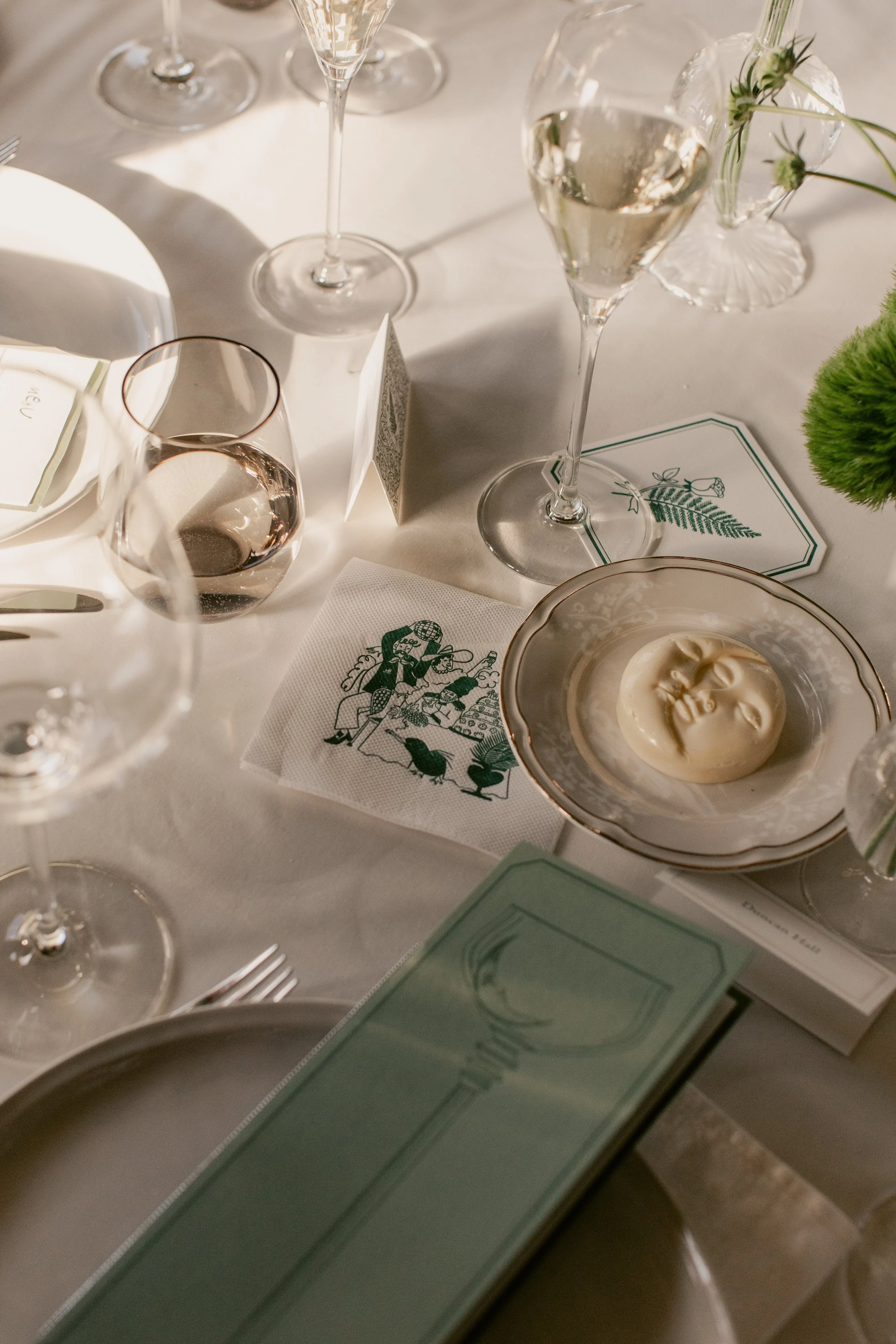 A table set for a meal with wine glasses, a water glass, a bowl of creamy dip, floral decoration, and printed menus or place cards, with soft natural lighting.