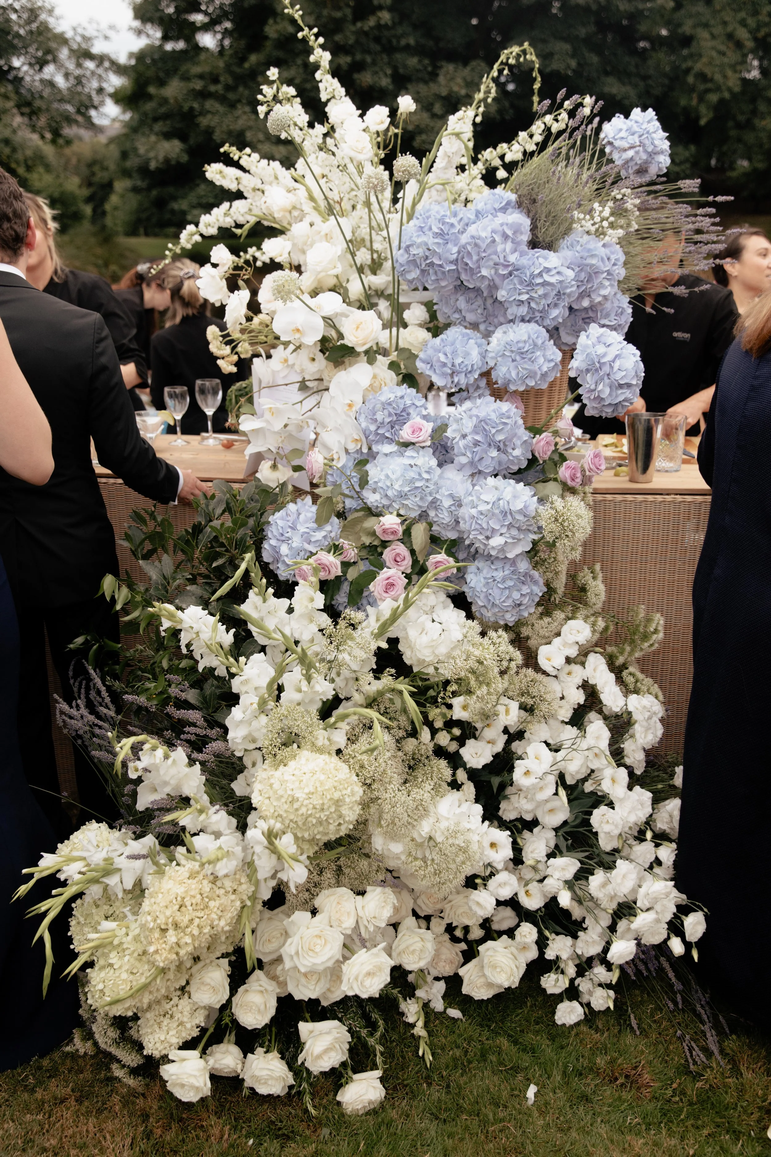 A large arrangement of white and light purple flowers, including hydrangeas, roses, and other blooms, set outdoors at a gathering or event, with people in formal attire in the background.