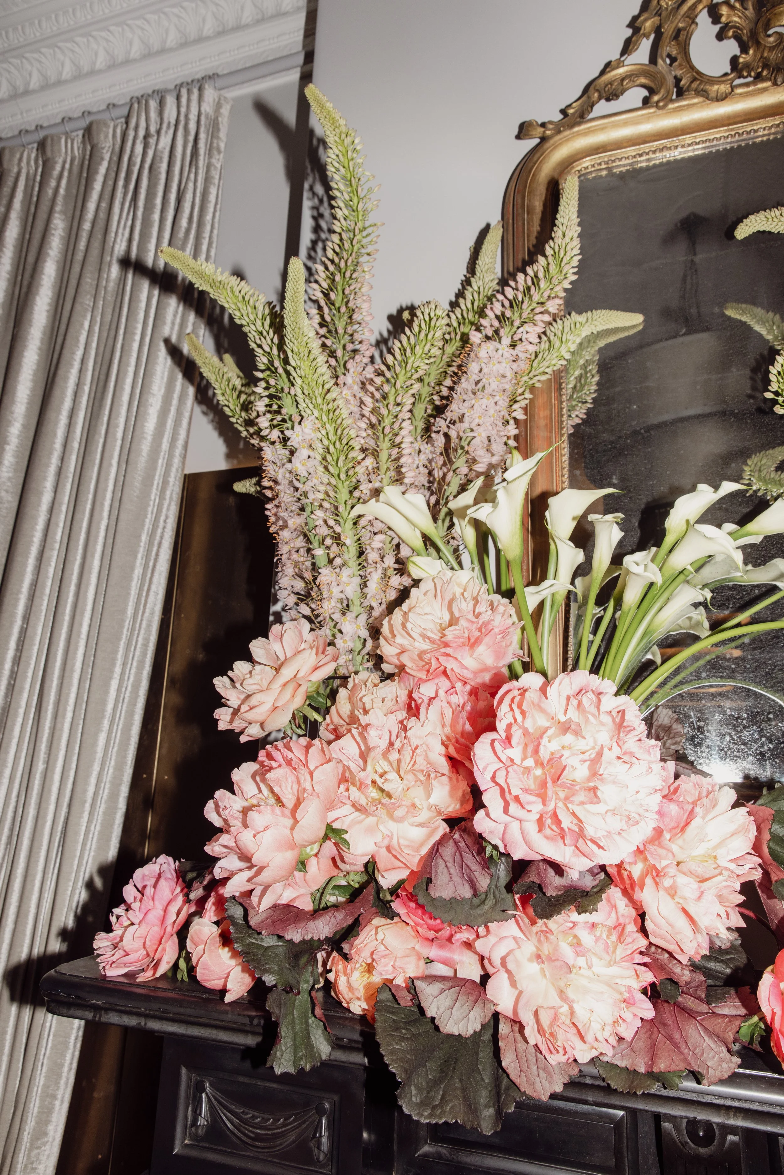 A floral arrangement featuring tall pink and green flowers, white calla lilies, and pink hydrangeas in front of a large mirror and a curtain.