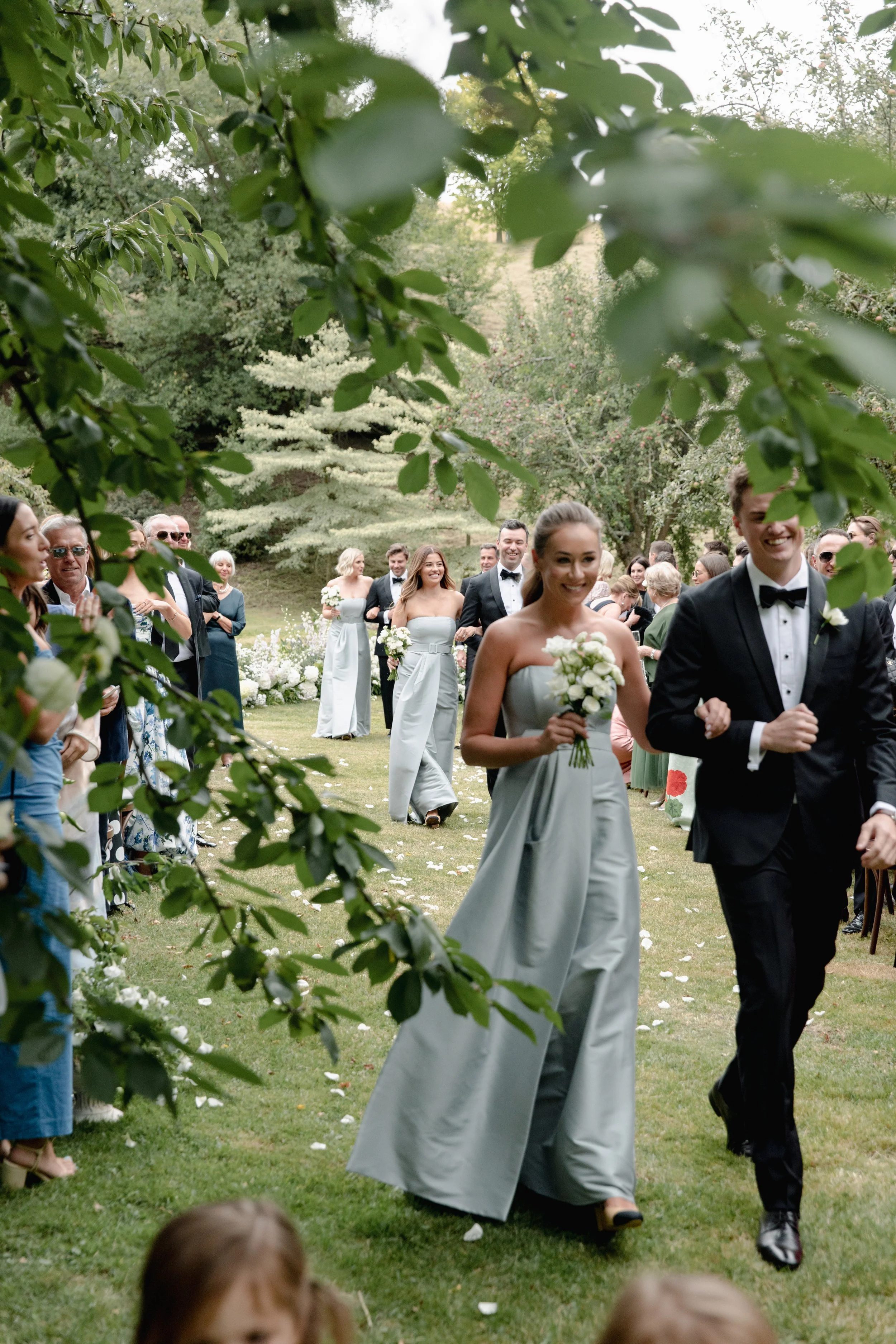 A wedding procession outdoors with bridesmaids in light gray dresses and groomsmen in black tuxedos, walking across a grassy area surrounded by trees and guests, with some people clapping and celebrating.