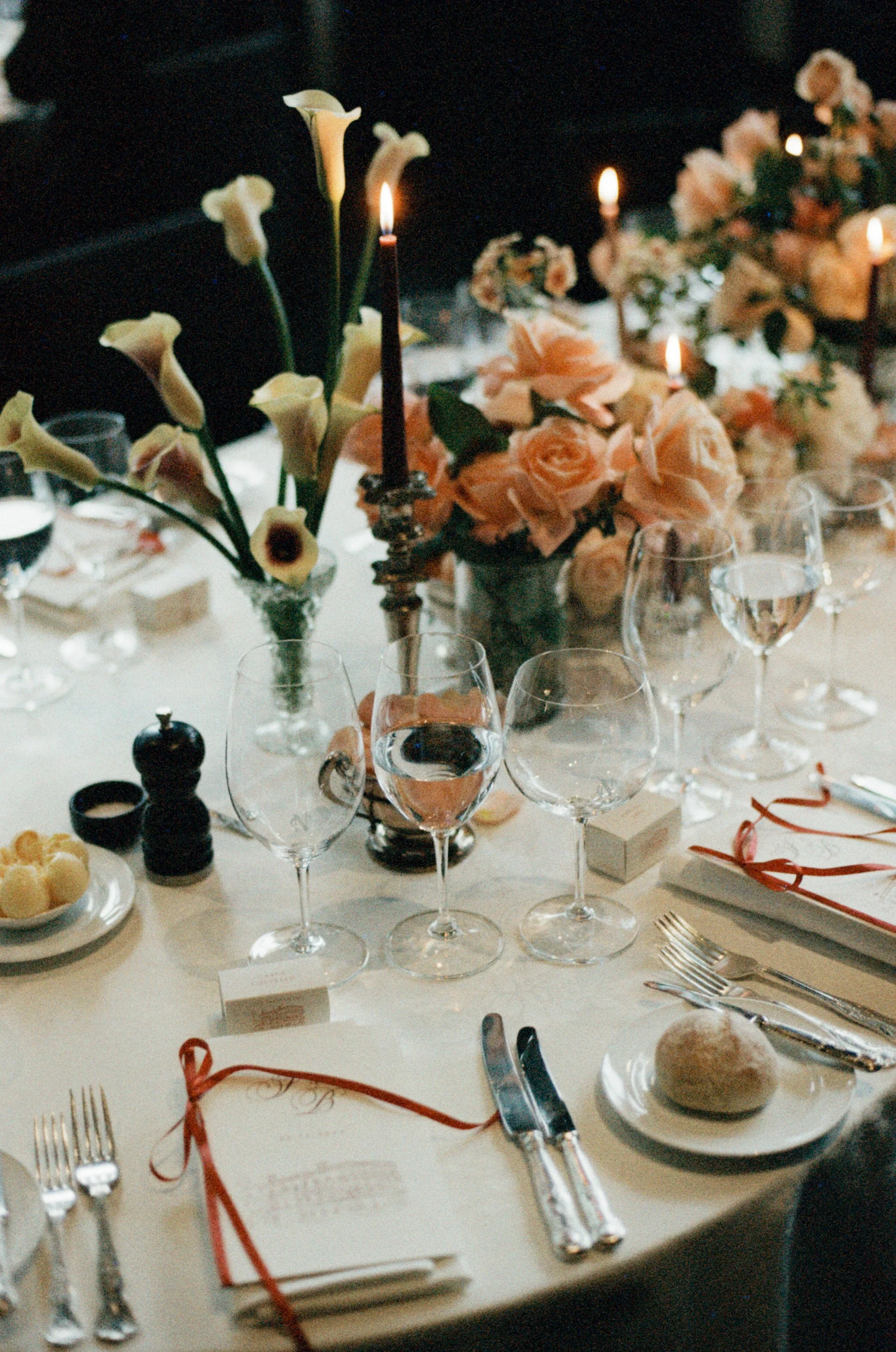 A formal dining table decorated with flowers, candles, wine glasses, plates, utensils, and small dessert treats, set for a special occasion or celebration.
