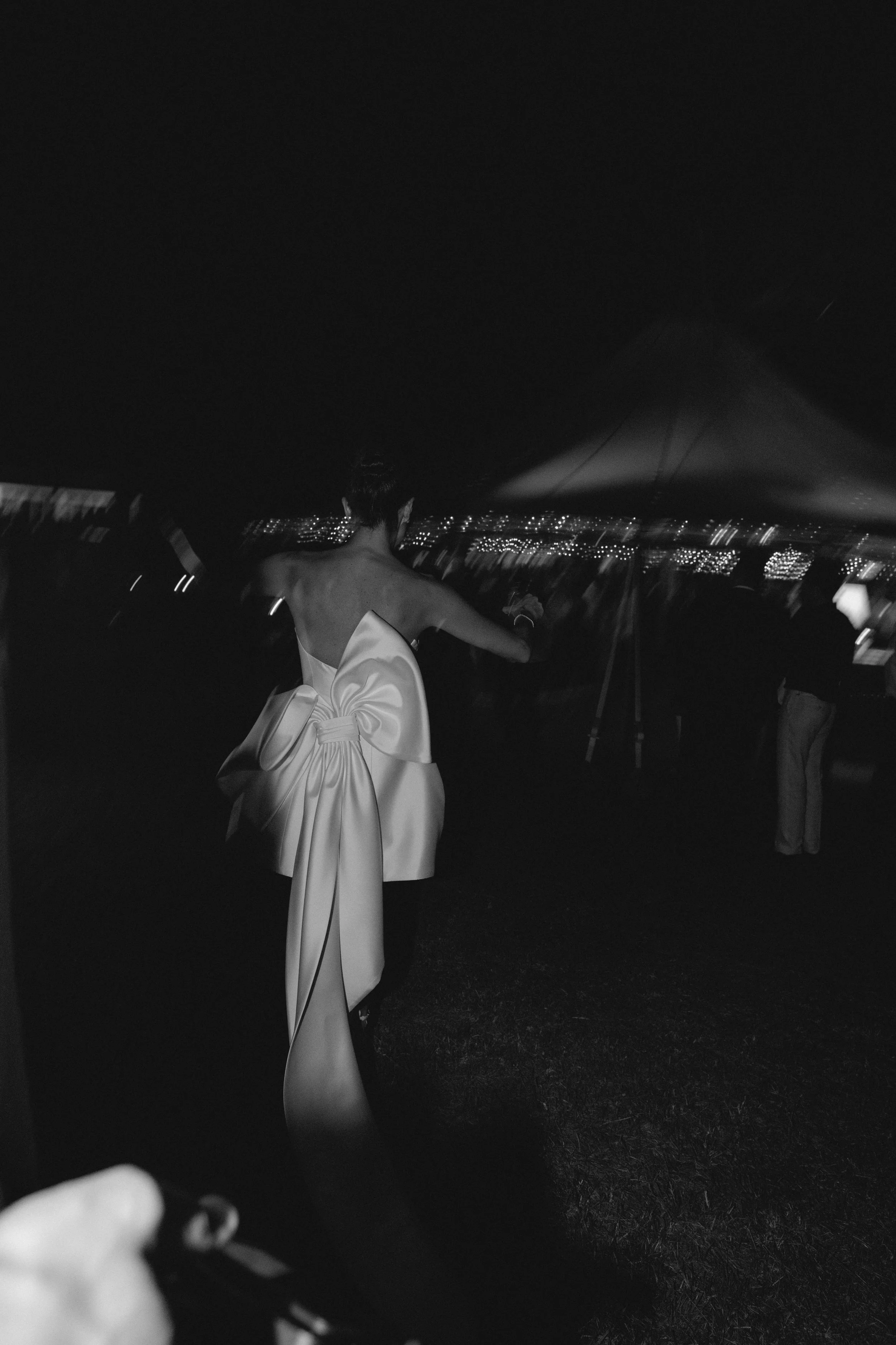 A woman in a light-colored gown with a large bow on her back is standing outdoors at night in front of city lights, with her back to the camera. A man on the right is also facing away, and a person's hand with a camera is visible in the foreground.