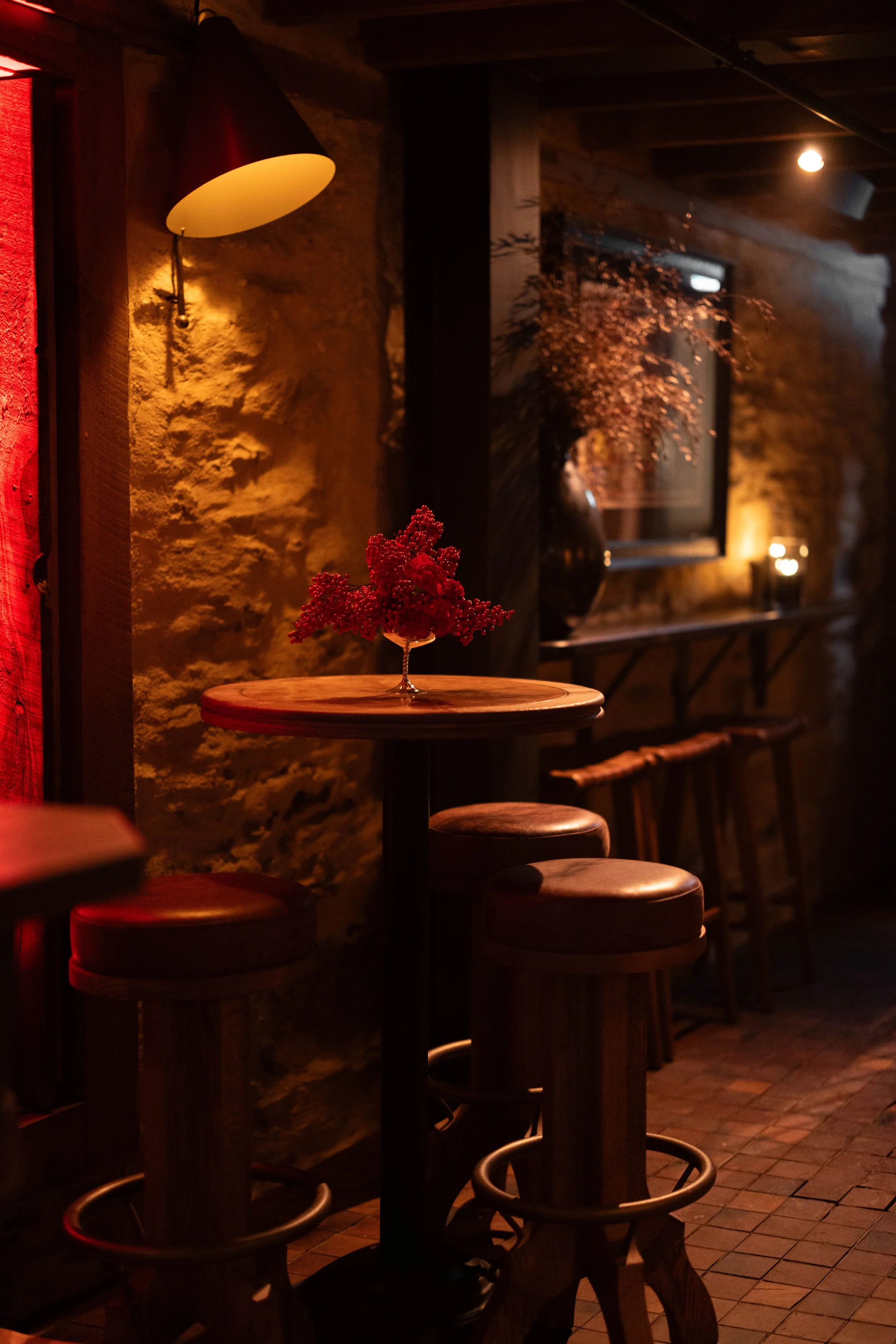 A cozy dimly lit bar or restaurant interior with wooden stools around high tables, a textured brick wall, and warm ambient lighting from wall sconces and candles, with decorative dried flowers on one table.