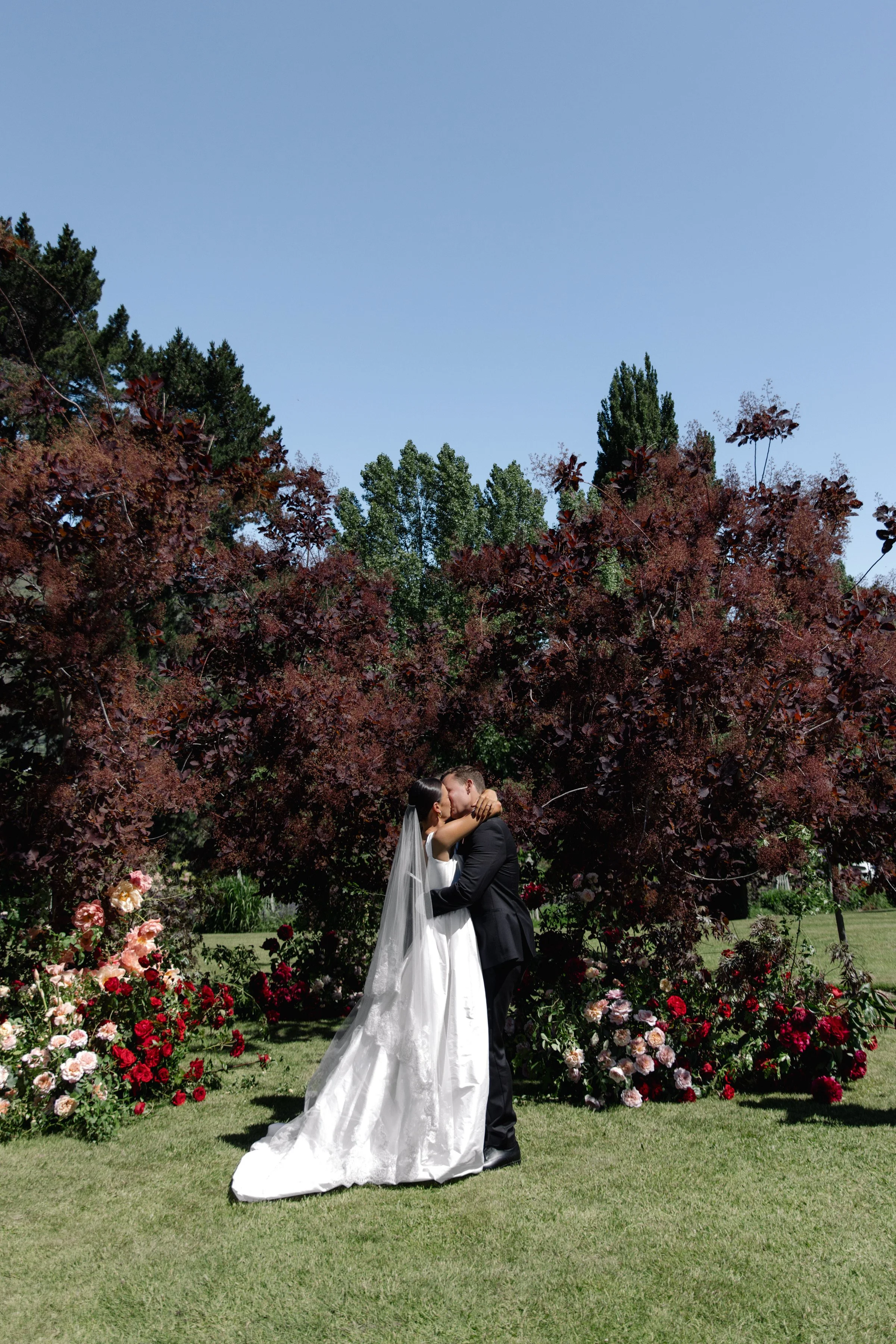 A bride and groom kissing outdoors under a large tree, surrounded by colorful flowers, on a sunny day.