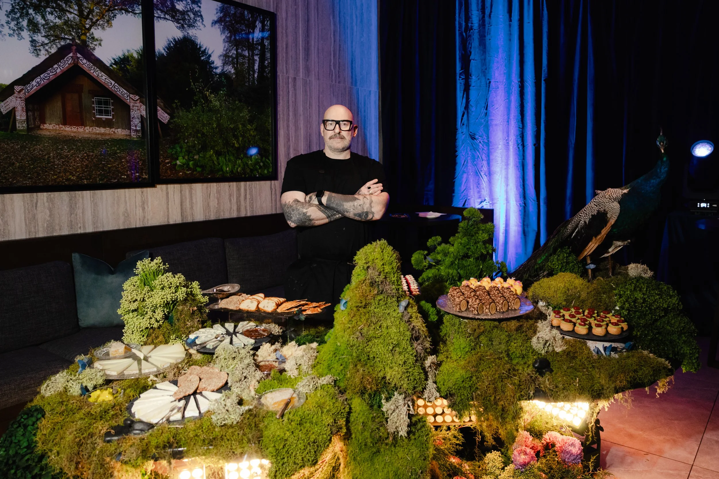 Indoor scene with a man standing behind a table of assorted desserts and decorations, with a large peacock statue, framed artwork of a house and trees on the wall, and dark curtains illuminated with blue light.