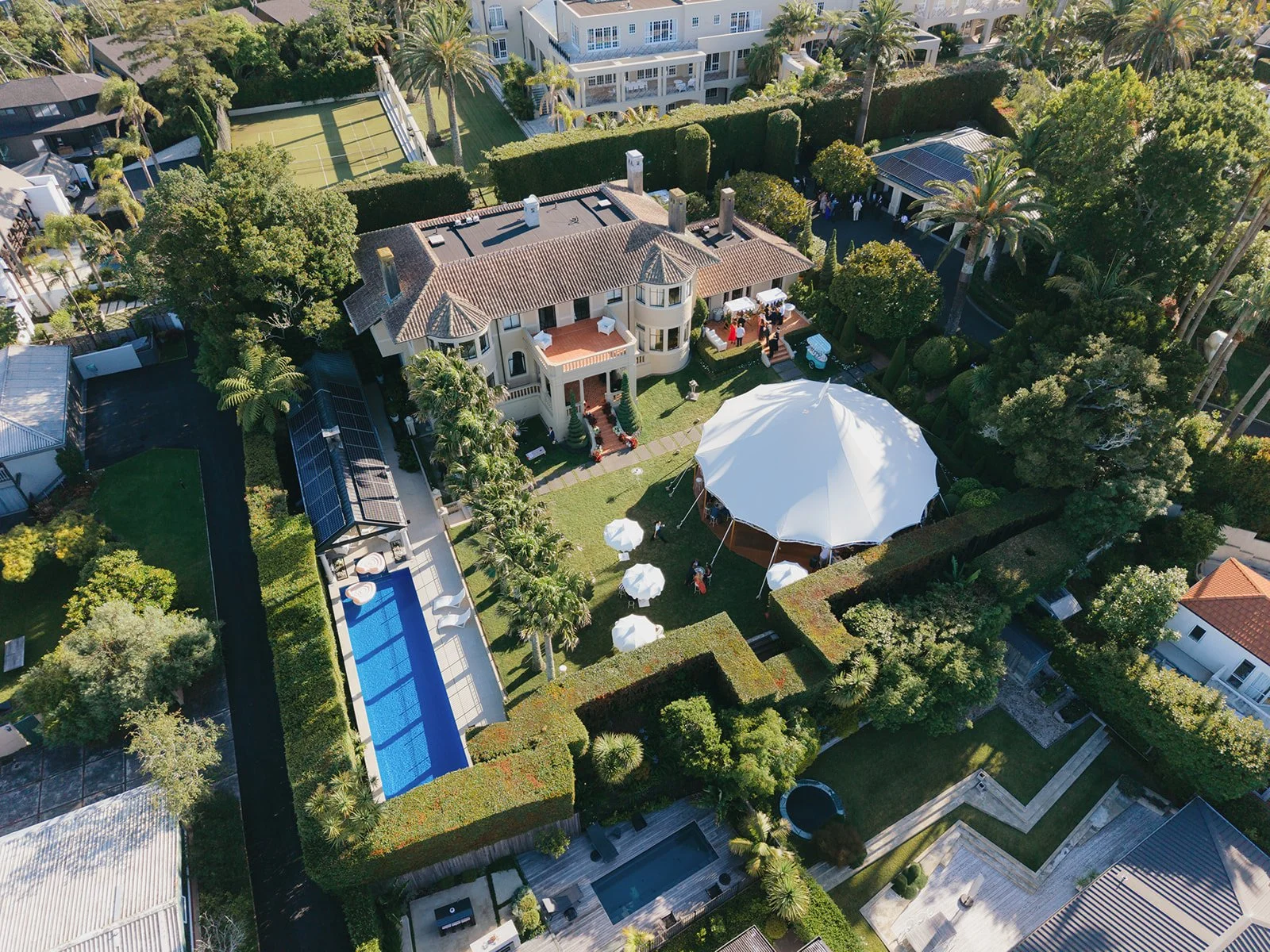 Aerial view of a large house with a red-tiled roof, surrounded by lush greenery, palm trees, and other foliage. The front yard features a lawn with white umbrellas and a large white canopy tent. There is a swimming pool with a covered area and a patio with outdoor furniture, and a tennis court is visible in the background.