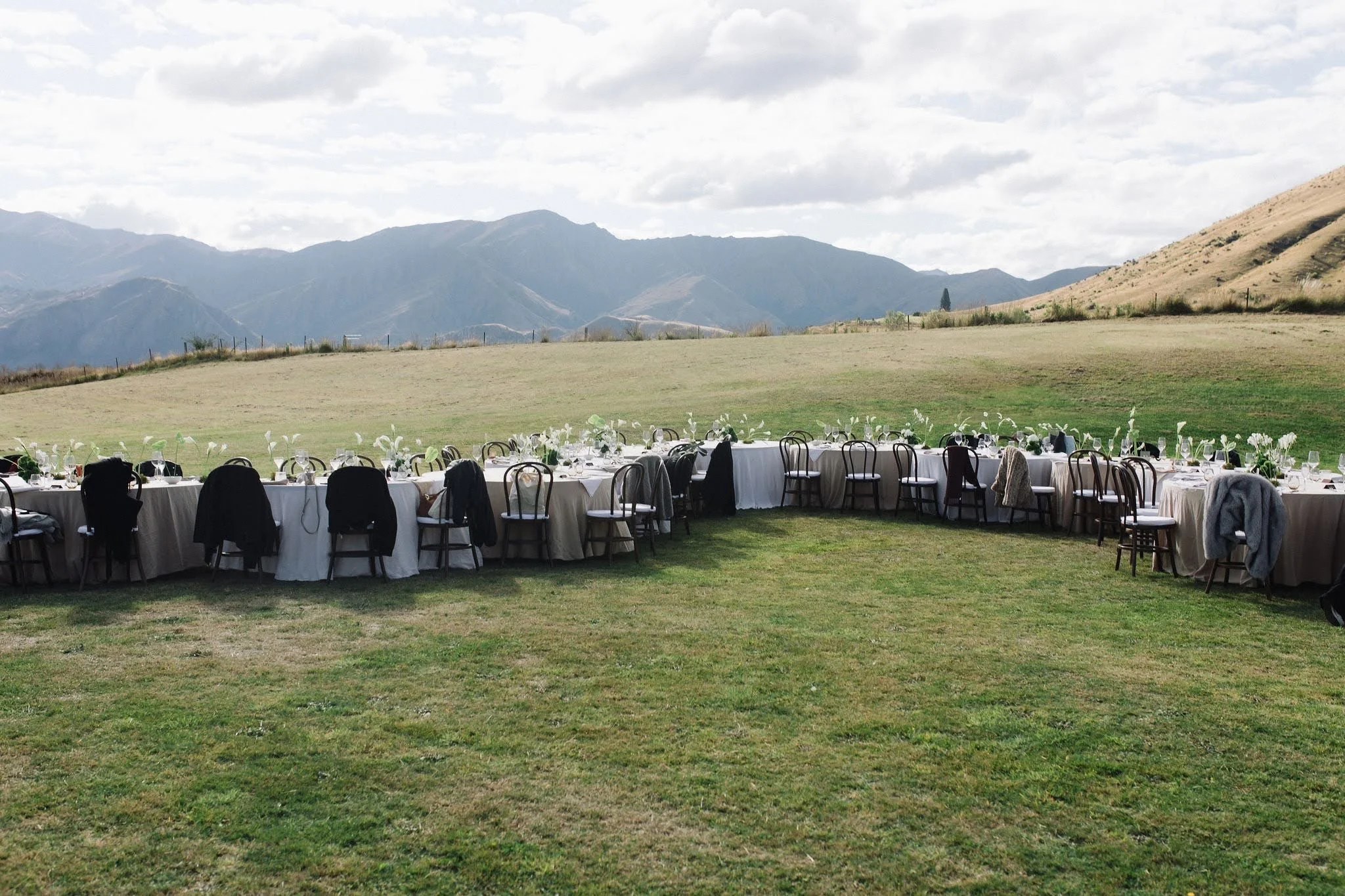 Outdoor dining setup with round tables covered in white tablecloths and black chairs, arranged on a grassy field with mountains in the background under a partly cloudy sky.