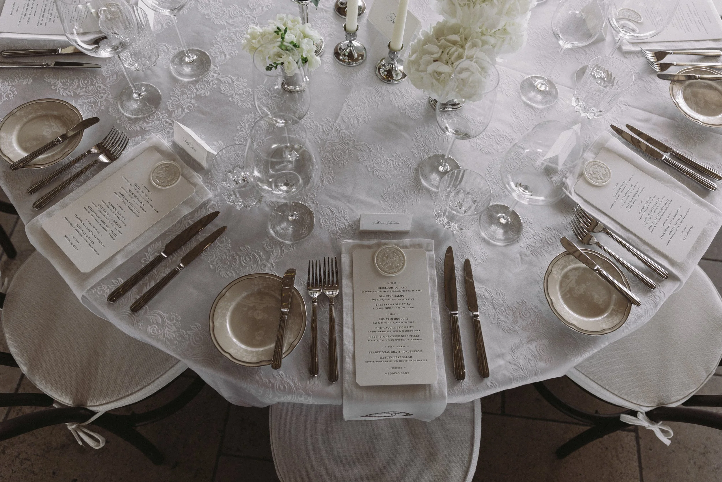 Round banquet table dressed in white tablecloth with elegant dishware, glassware, and silverware arranged for a formal event. There are white floral centerpieces and menu cards placed in front of each setting.