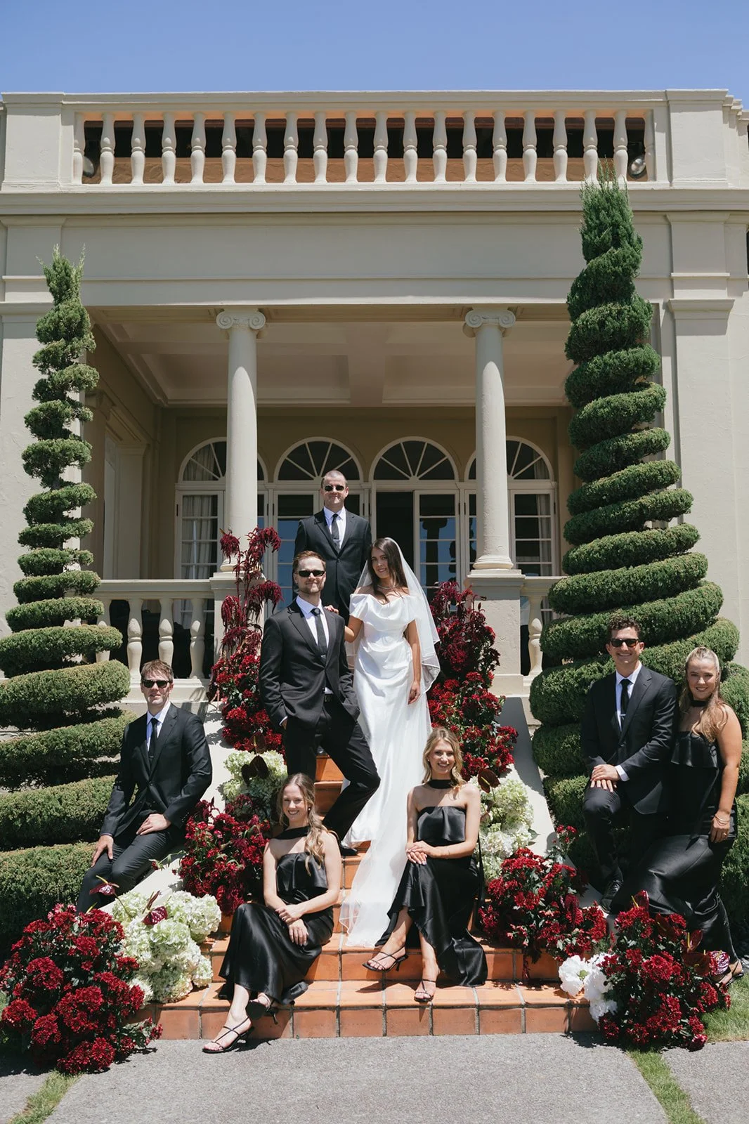 A wedding party with seven people, including a bride in a white dress and six friends in formal attire, standing on stairs decorated with red, white, and burgundy flowers in front of a large house with columns and topiary trees.