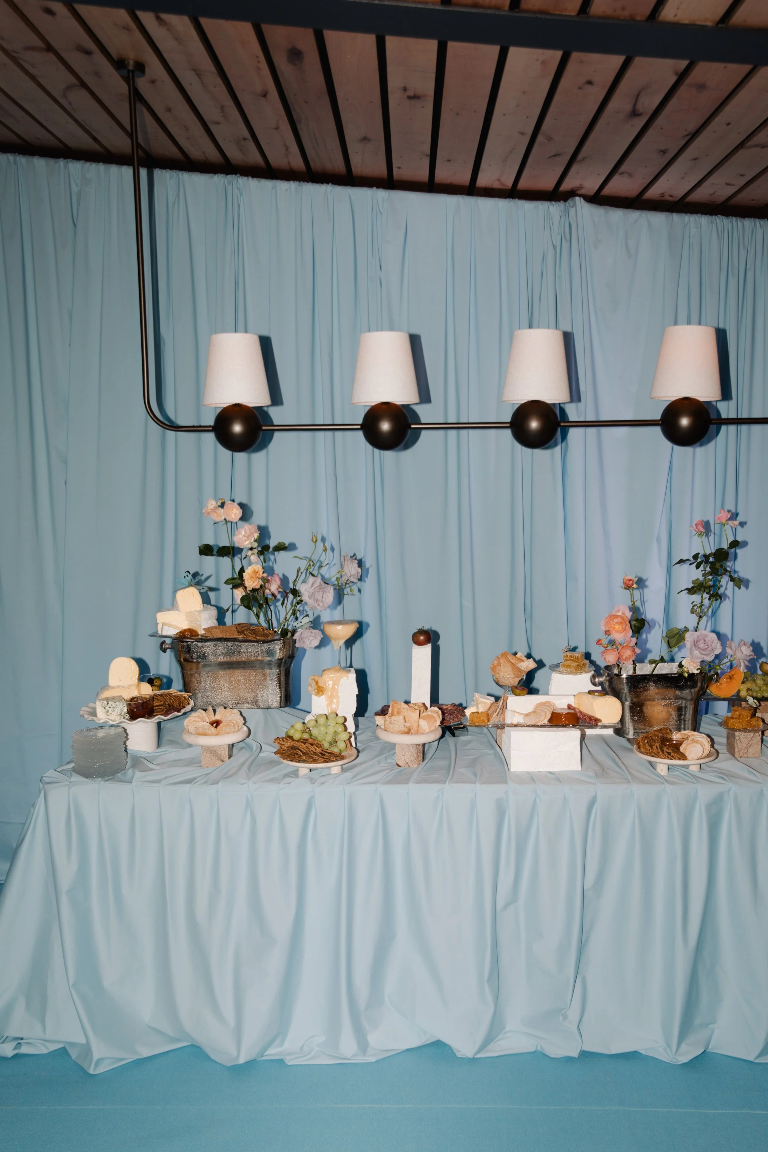 A buffet table covered with a blue tablecloth, featuring various cheeses, fruits, and snacks, set against a light blue curtain backdrop with a black and beige wall lamp overhead.