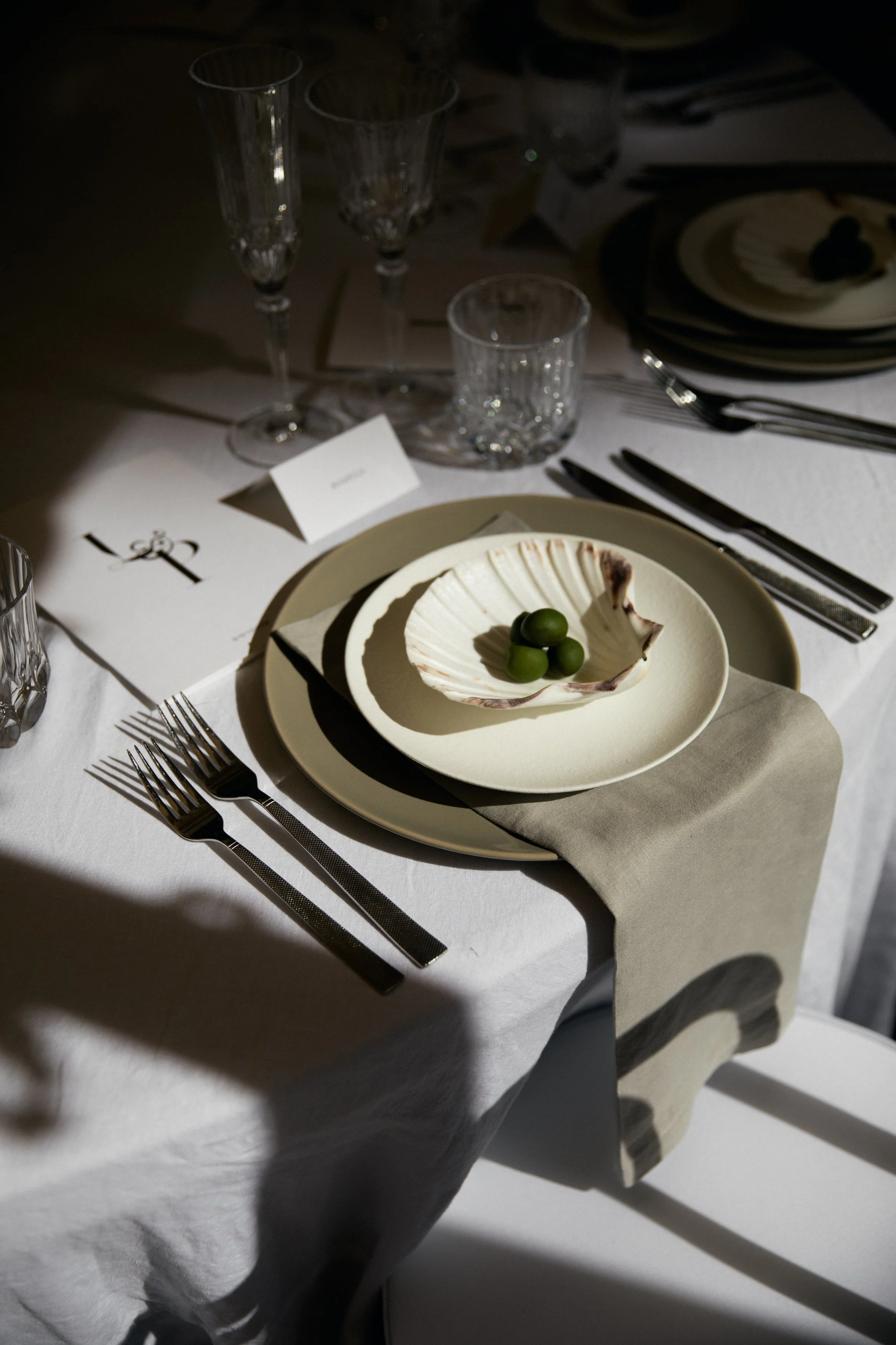 A formal dining table set with wine glasses, crystal tumblers, plates, and cutlery. A small bowl with green olives sits on a larger plate. The setting is illuminated with a spotlight, creating shadows on the white tablecloth and a beige napkin.